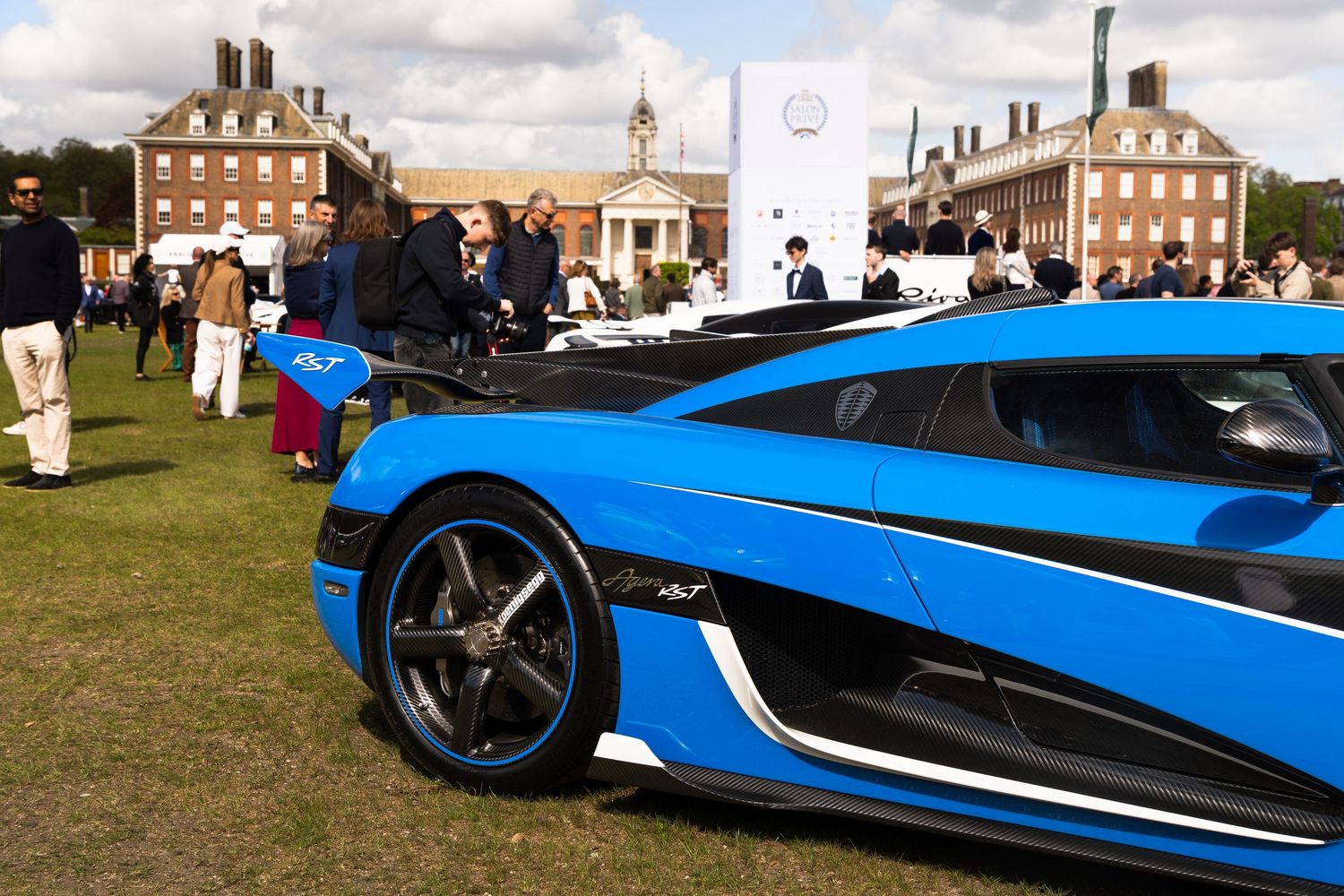 Blue Koenigsegg supercar on display at an outdoor luxury car show with crowds and historic buildings.