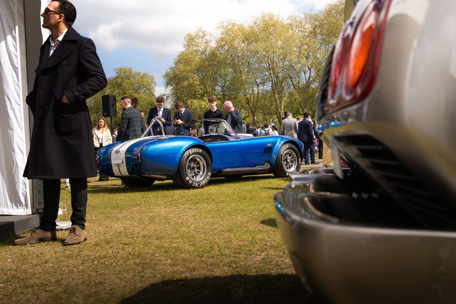 A classic blue AC Cobra roadster on display at an outdoor luxury car show with well-dressed attendees.
