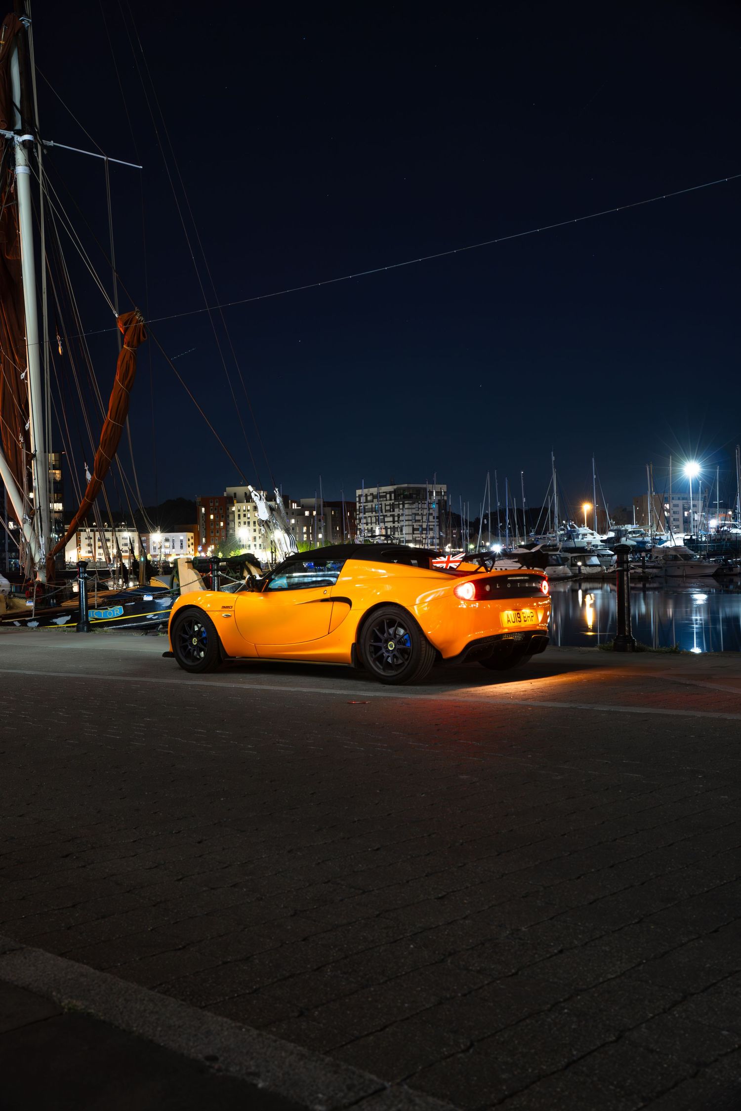 Rear view of a yellow Lotus Elise at a marina at night with sailboats and glowing lights.