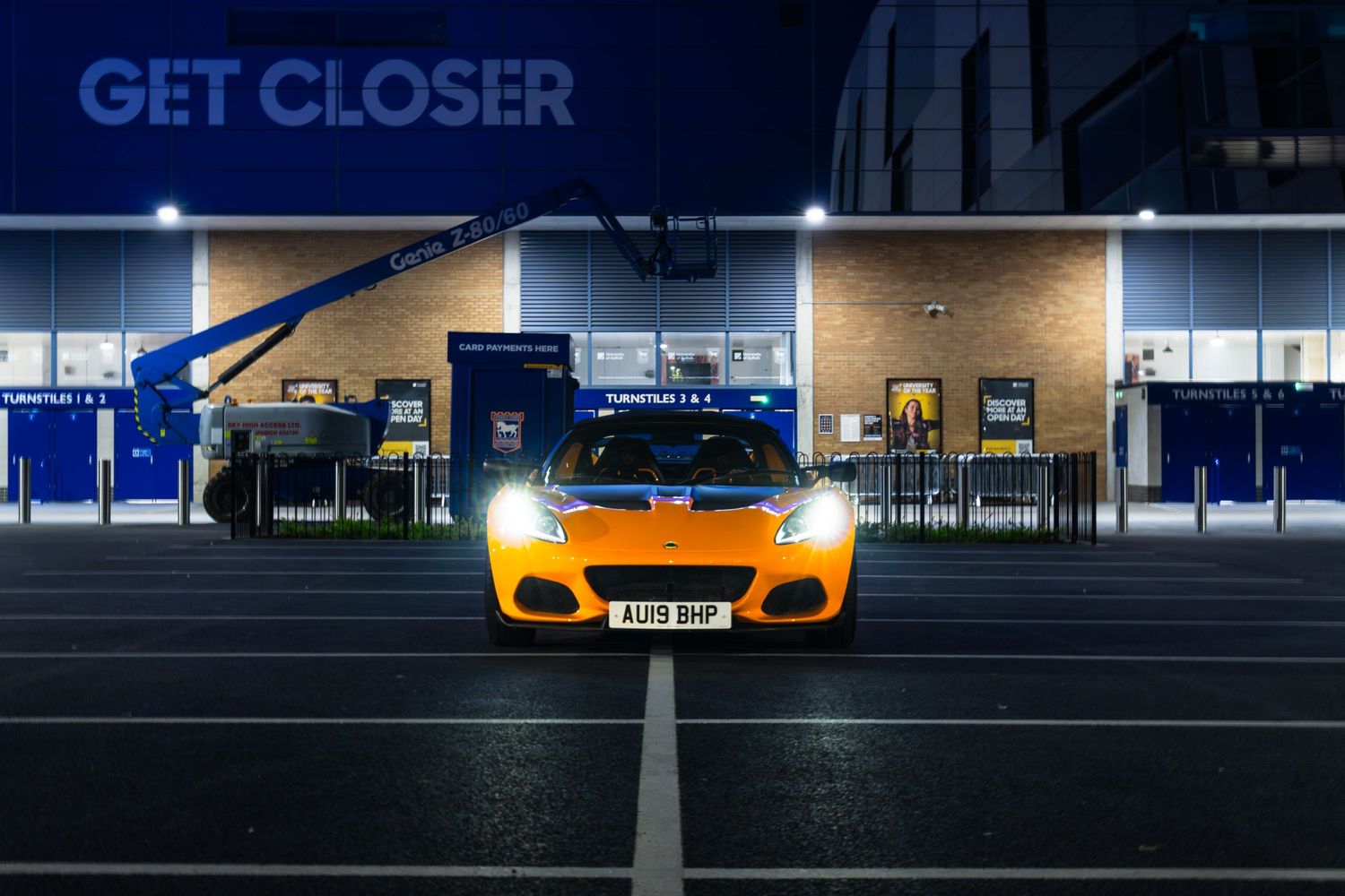 Front view of orange Lotus Elise with registration AU19 BMP outside GET CLOSER stadium at night.