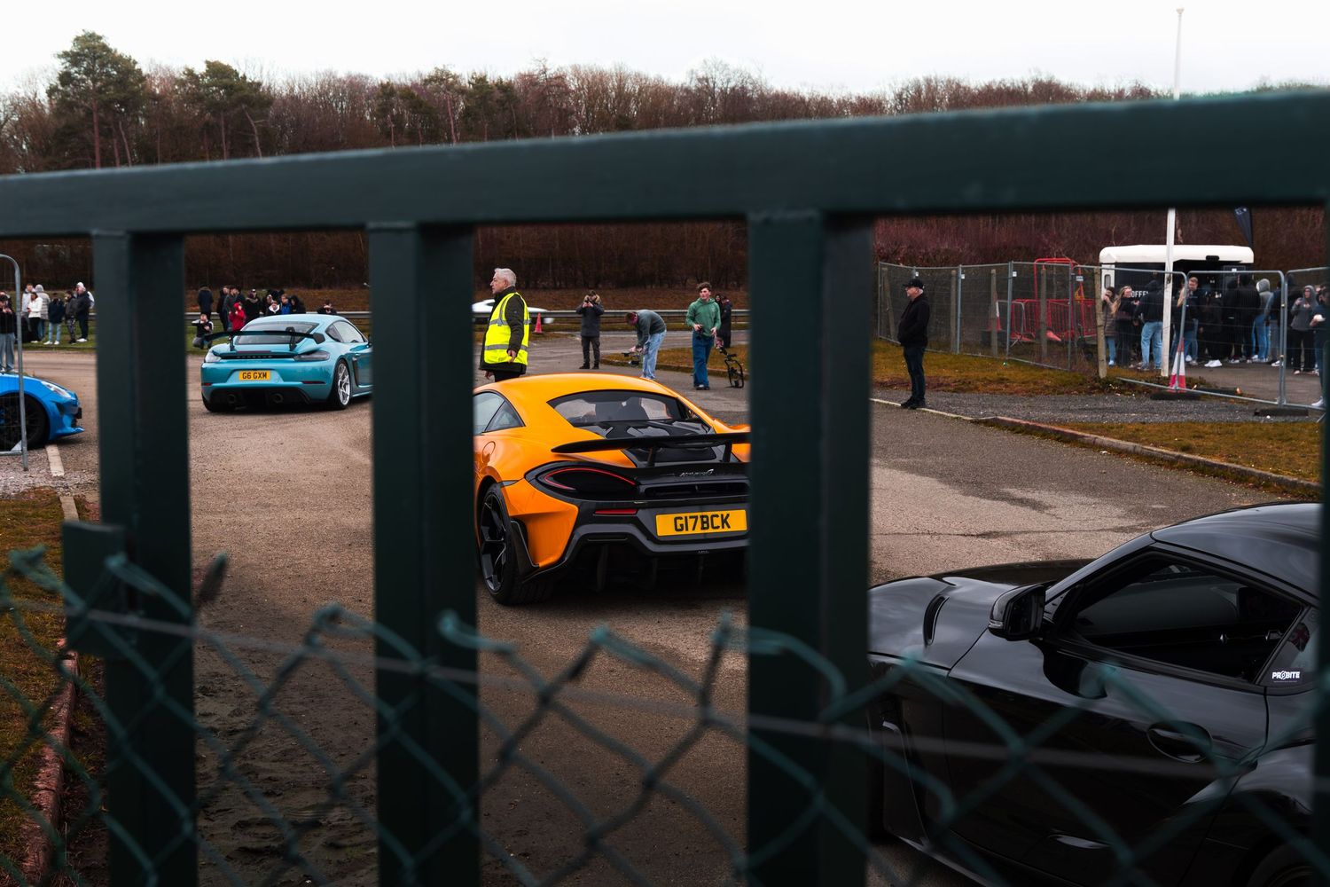 Orange McLaren supercar viewed through chain-link fence at an outdoor car meet event.