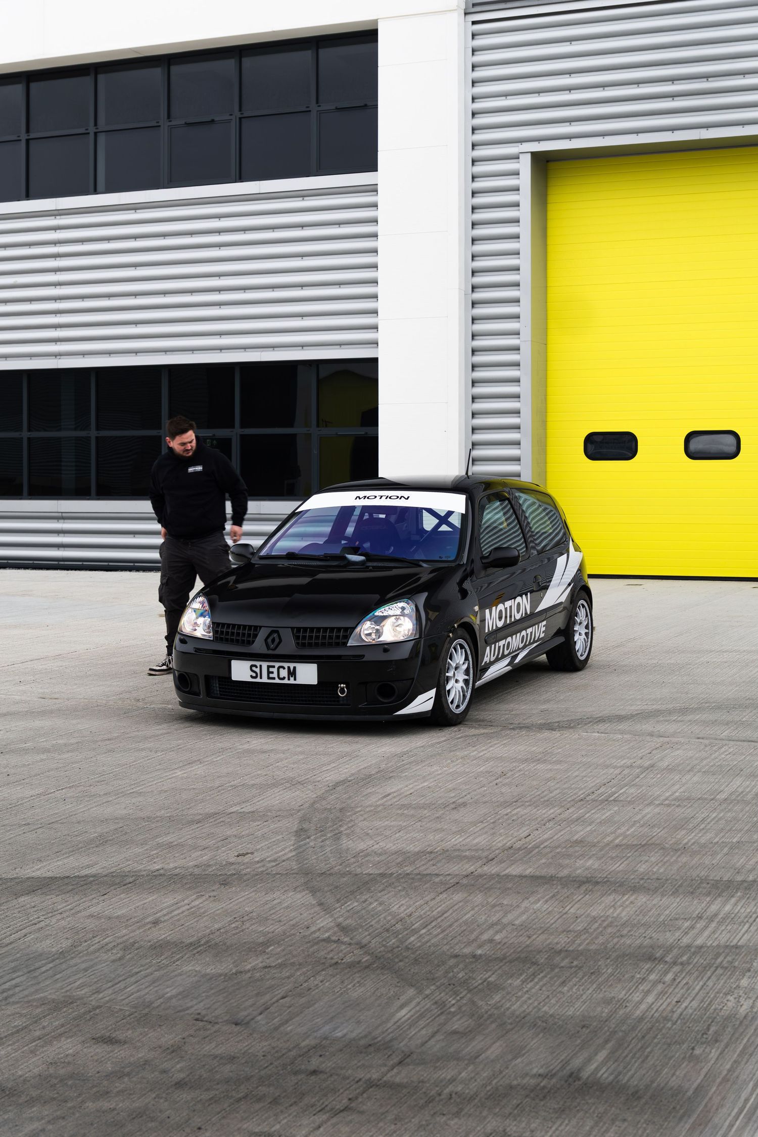 Man inspecting a black Renault Clio race car with motorsport livery parked outside yellow garage doors.