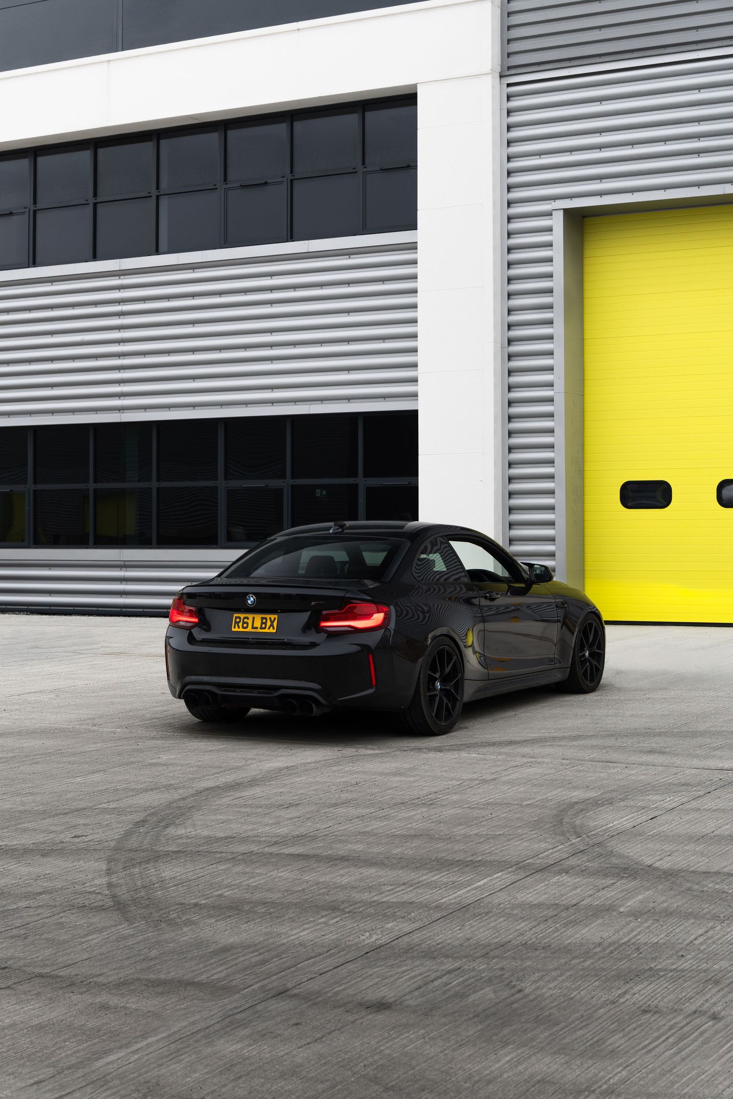Black BMW M2 coupe parked on marked concrete outside a yellow-doored industrial unit.