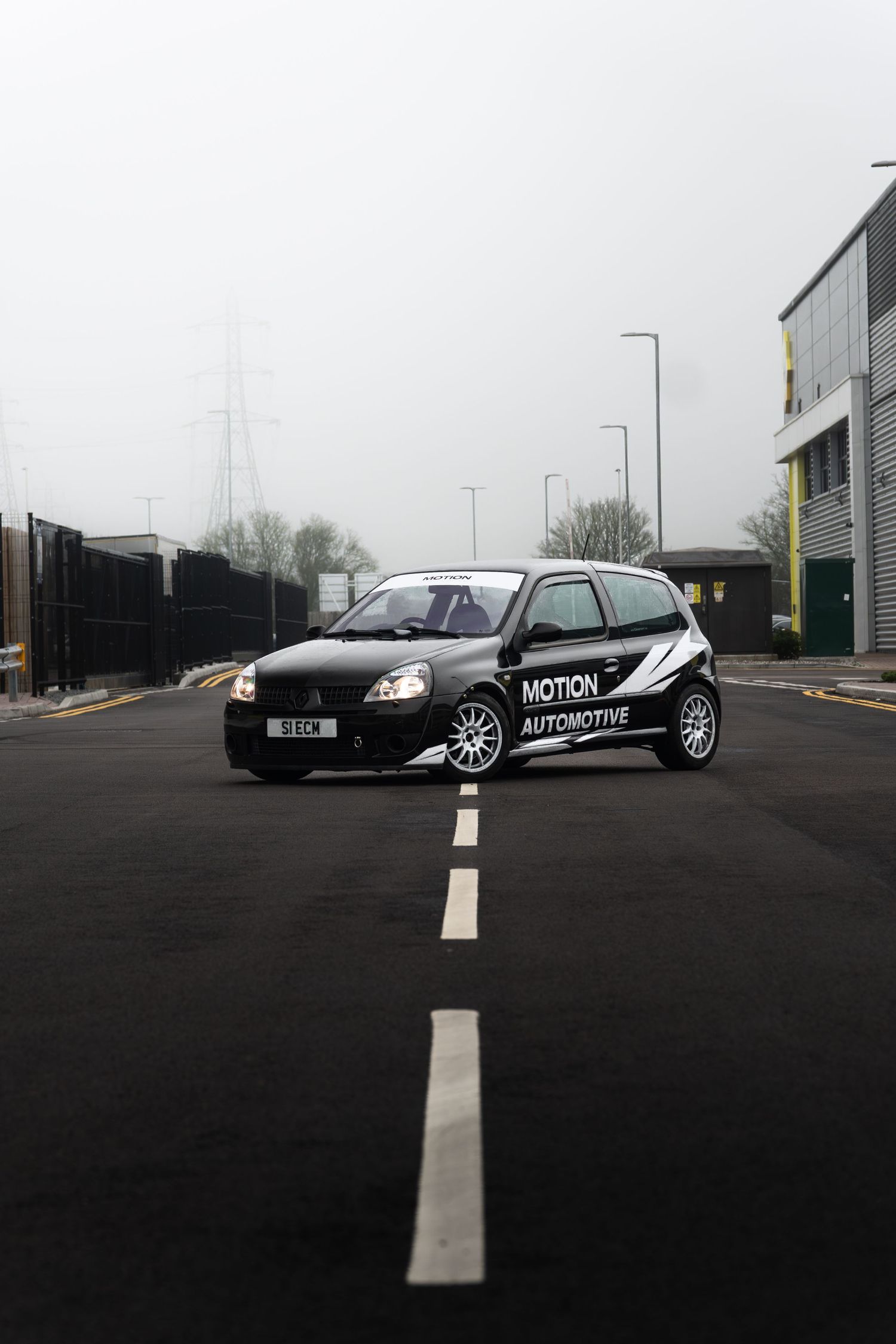 Black Motion Automotive Renault Clio with headlights on positioned on a misty industrial estate road with white markings.