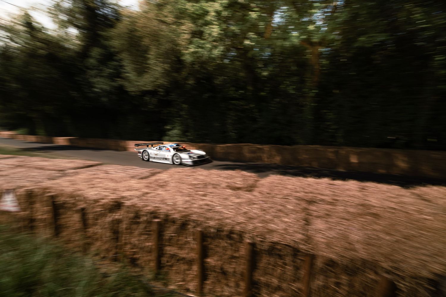 White race car speeding past hay bales on a tree-lined hillclimb course, captured with motion blur effect.