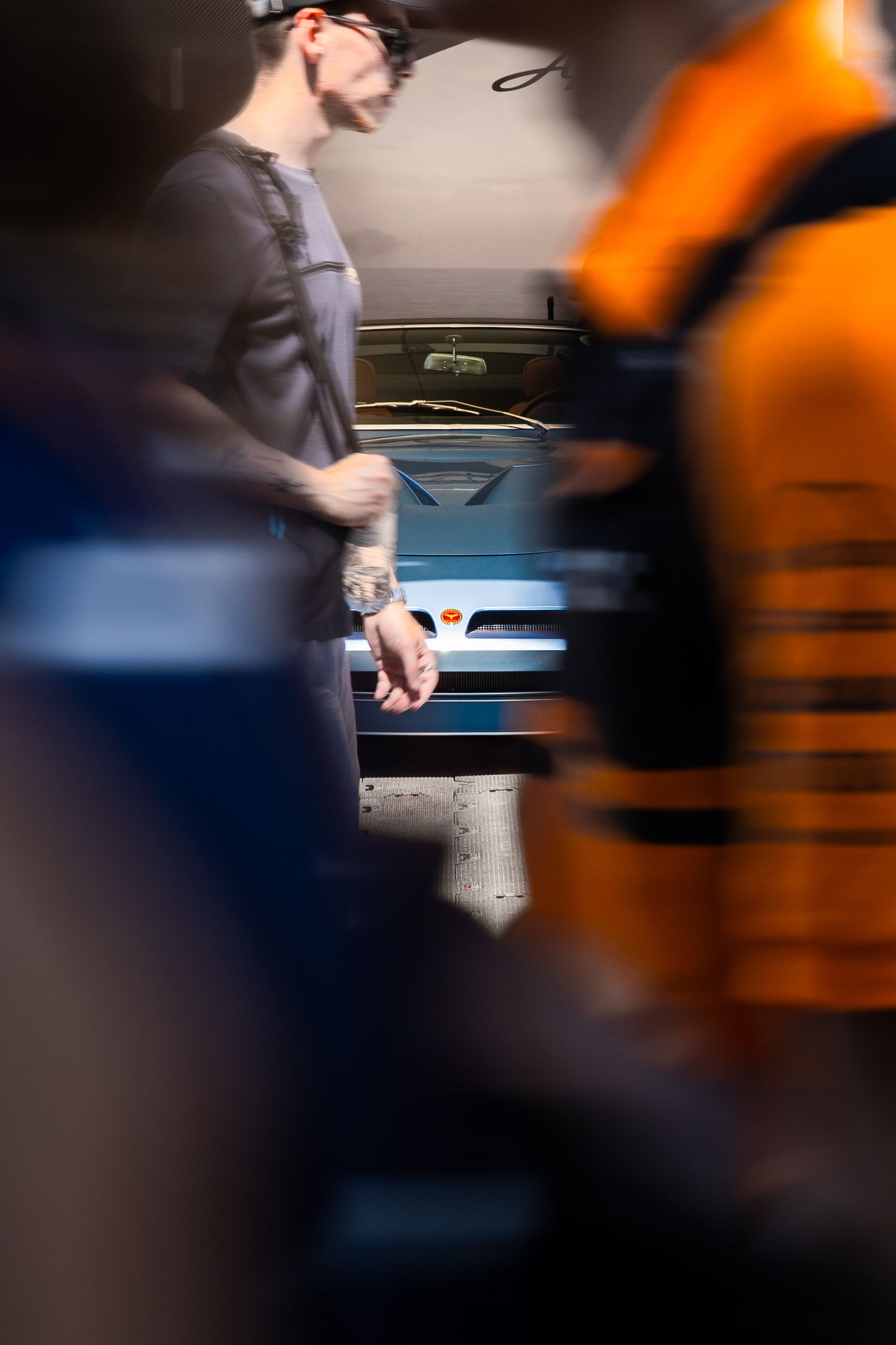 A mechanic in gray overalls stands in an auto repair shop beside a silver car, with blurred orange equipment in the foreground.