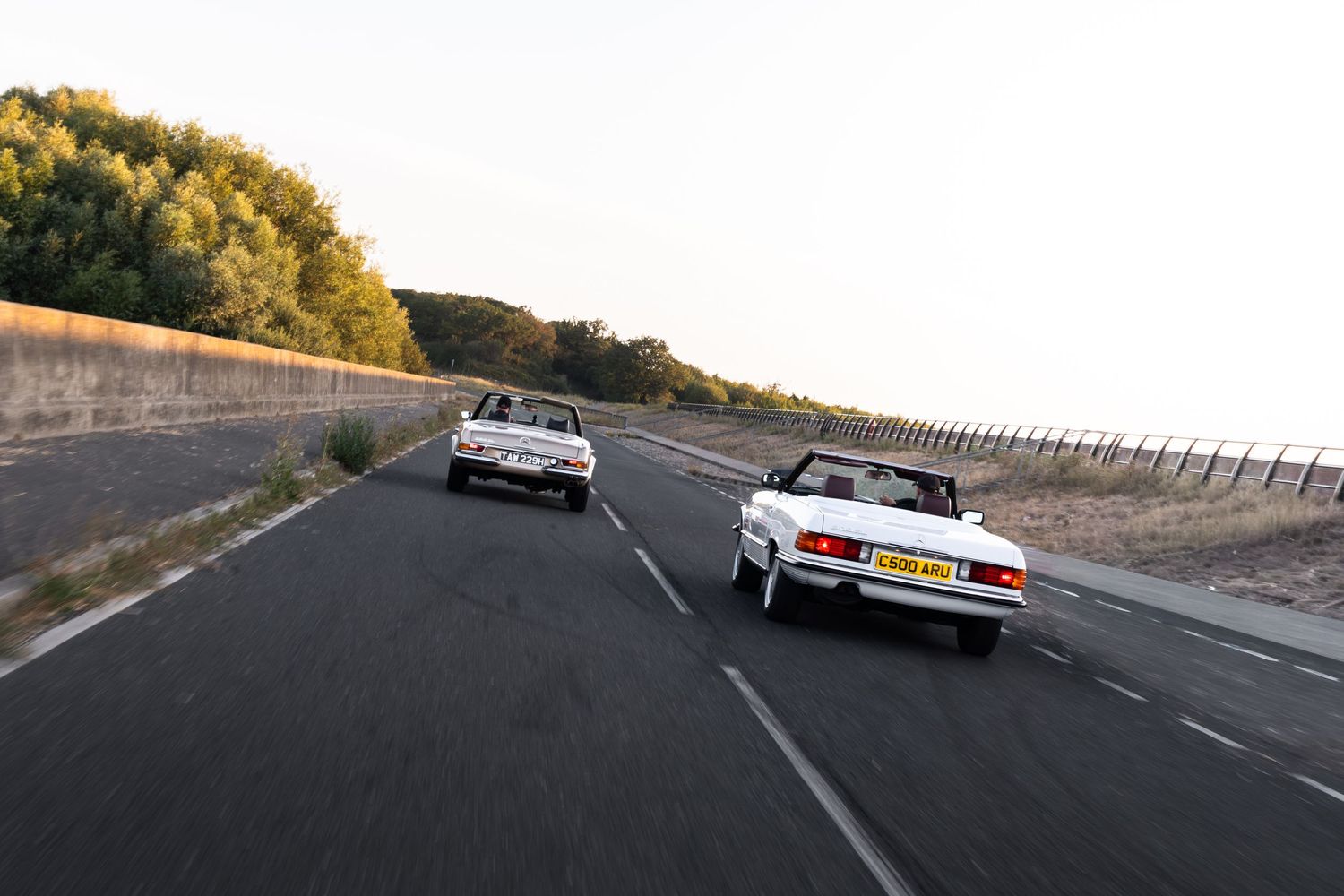 Two white convertible cars driving on an open road at sunset with trees and a stone wall in the background.