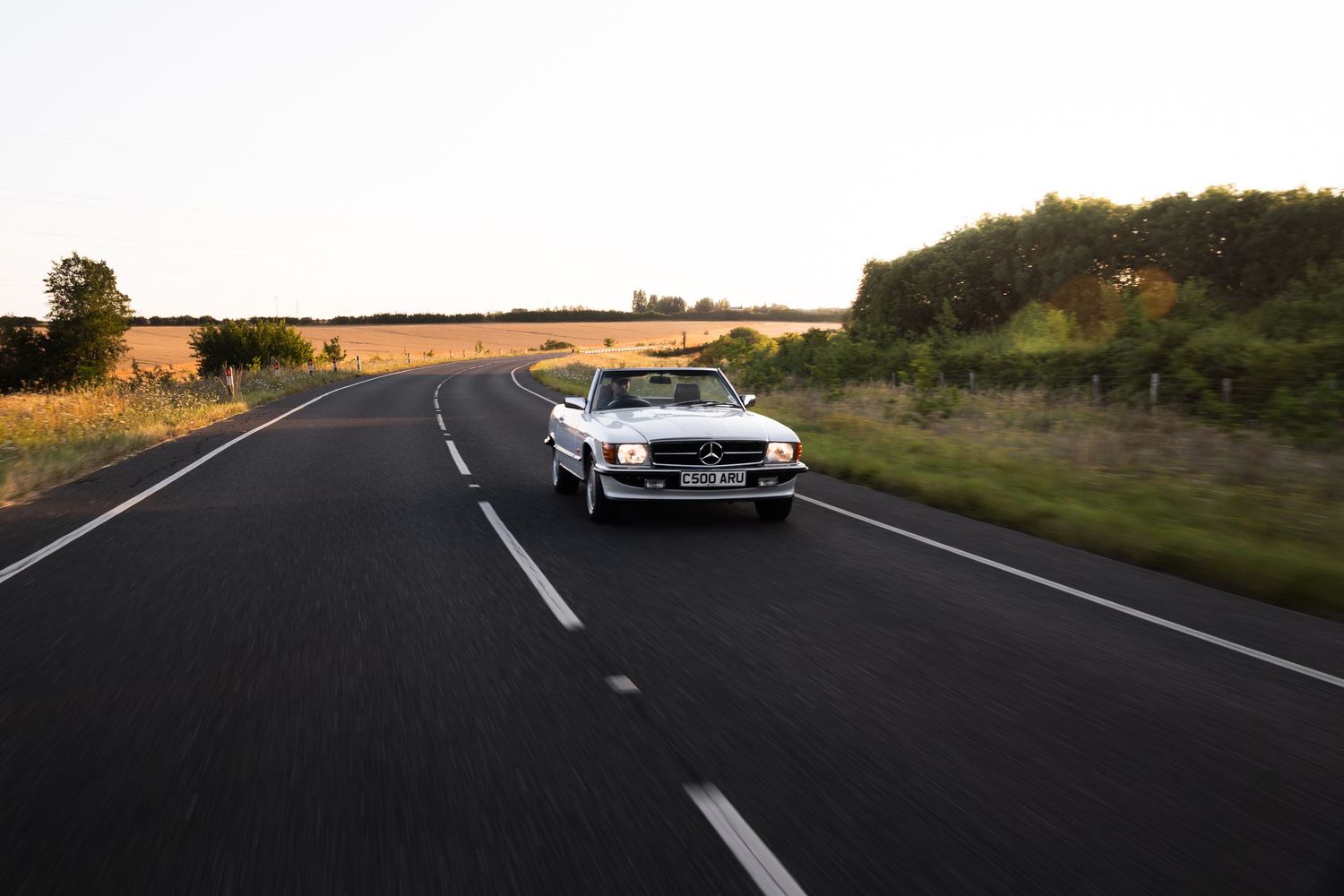 White Mercedes-Benz convertible driving on a scenic open road surrounded by green fields and trees at golden hour.