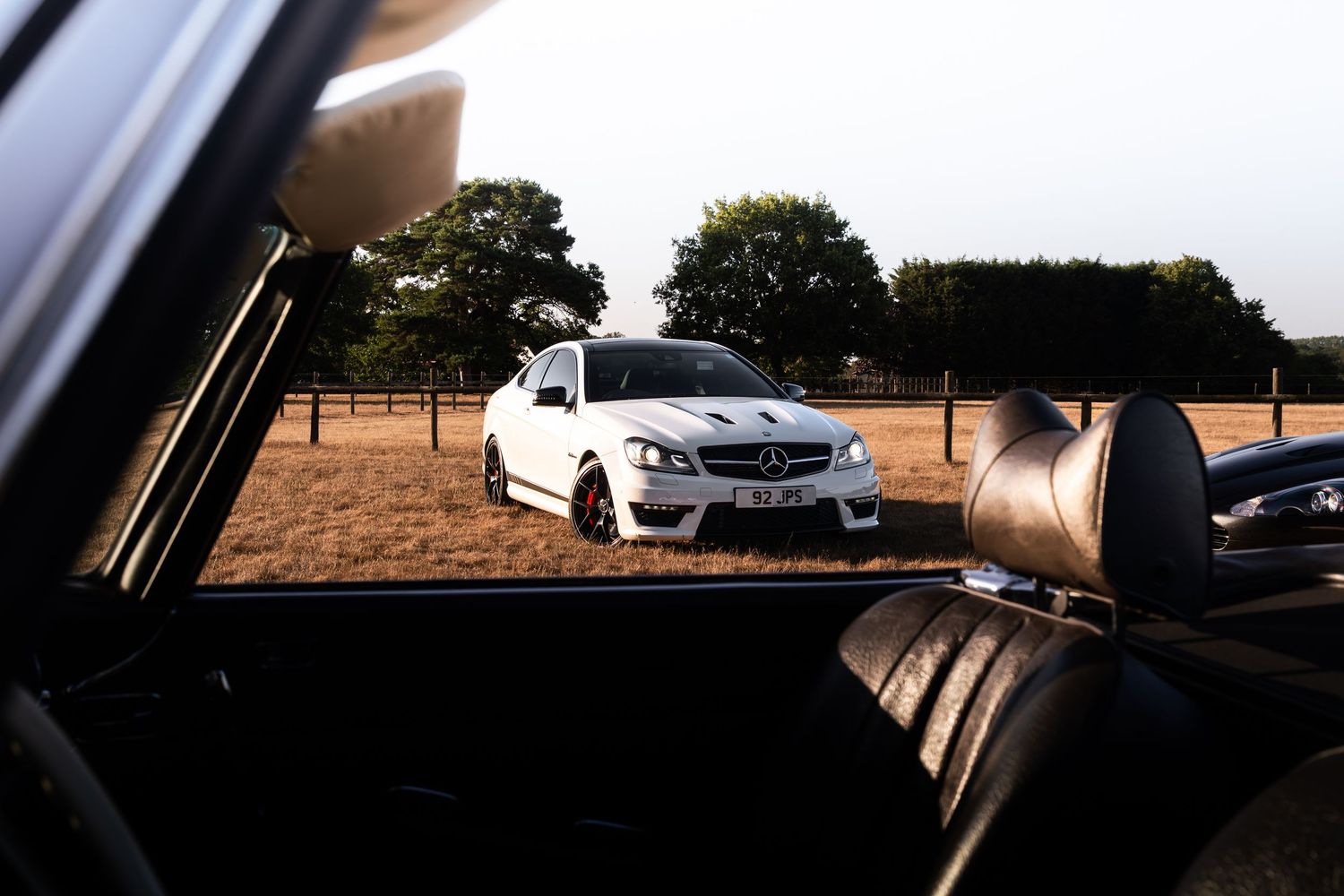 White Mercedes-Benz AMG C63 parked in a golden field, photographed from inside a classic black leather interior car.