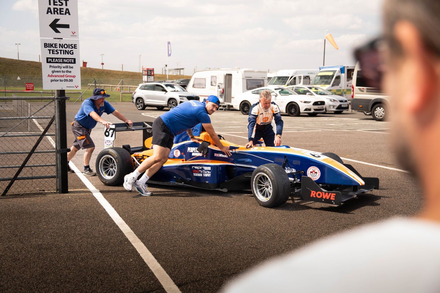 Pit crew members push a blue Formula racing car through the pit lane at a motorsport circuit.