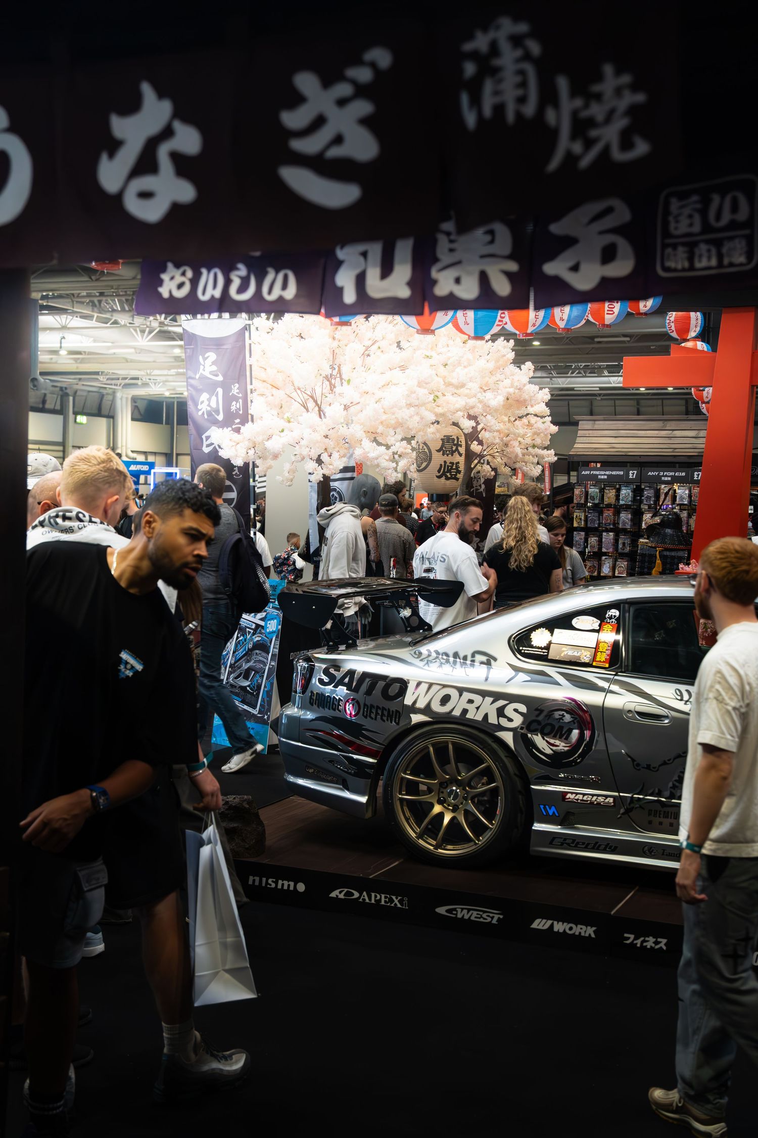 Modified drift car with Saitoworks livery displayed at Japanese-themed auto show with cherry blossom decor.