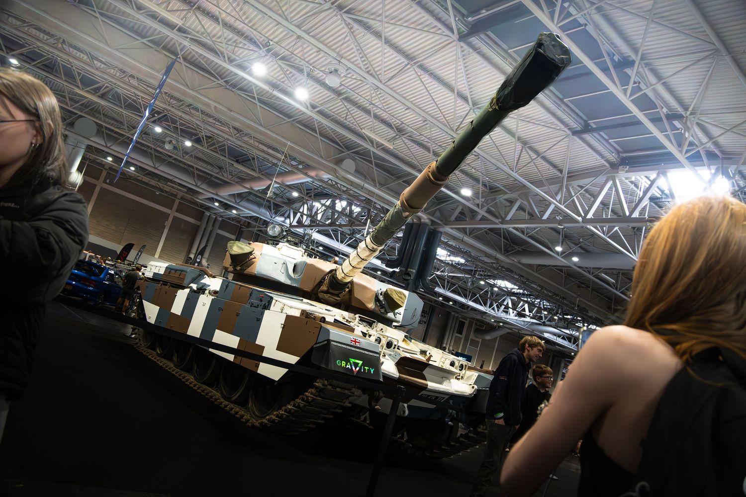 Military tank on display inside a large exhibition hall with visitors viewing it up close.