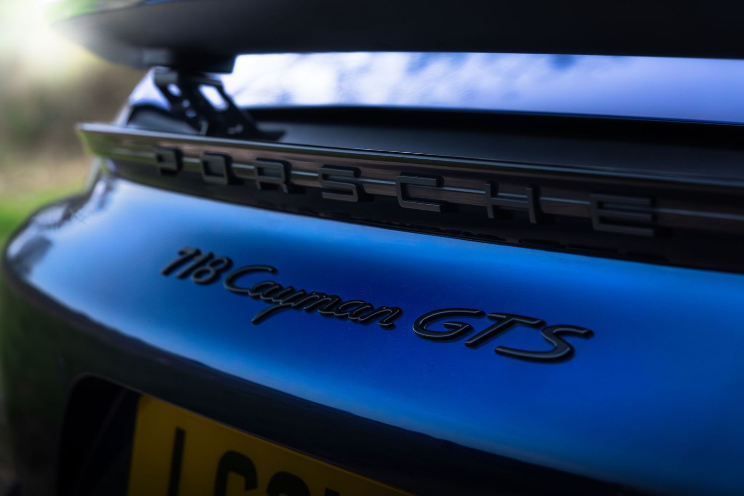 Close-up of Porsche 718 Cayman GTS rear badge and spoiler with UK number plate visible below.