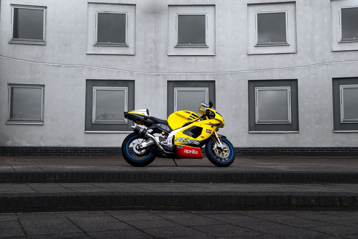 Yellow Aprilia RSV1000 sportbike parked on tiered steps against a minimalist gray building wall.