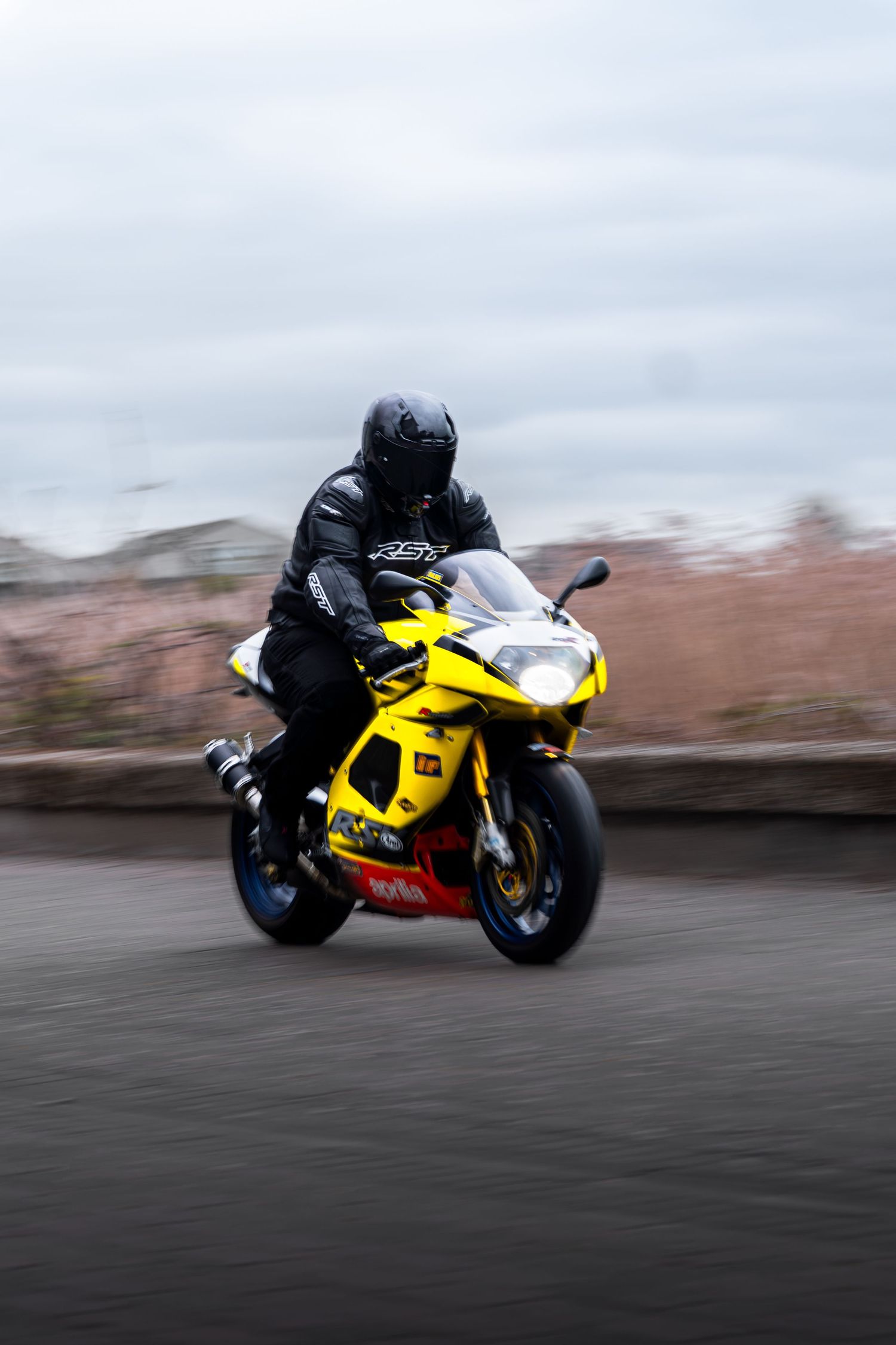 Motion-blurred yellow Aprilia RSV motorcycle with black-suited rider speeding on a road.