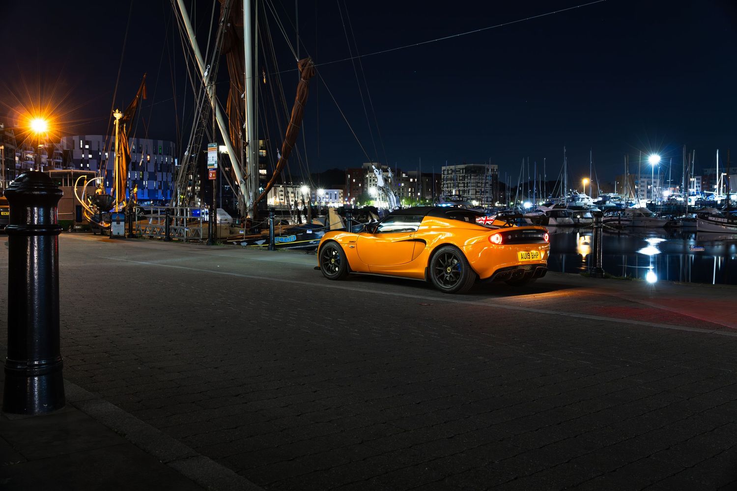 Yellow Lotus Elise parked beside tall sailboat masts at a marina during nighttime photography.