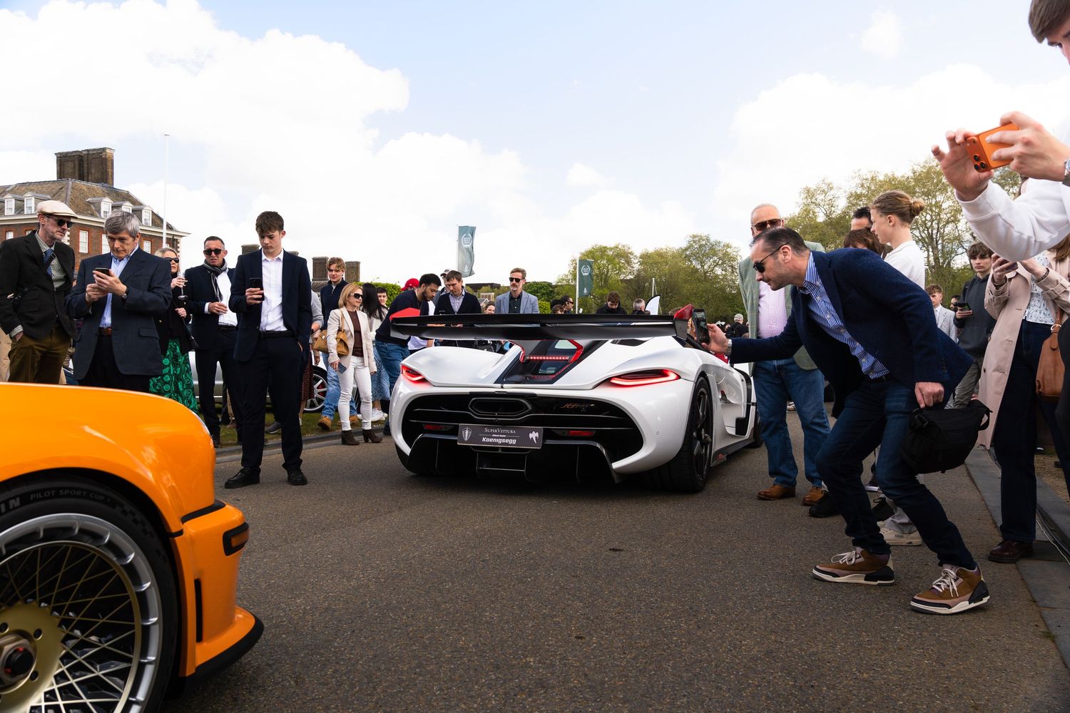 Crowd of spectators photographing a white Koenigsegg hypercar at an outdoor automotive show.