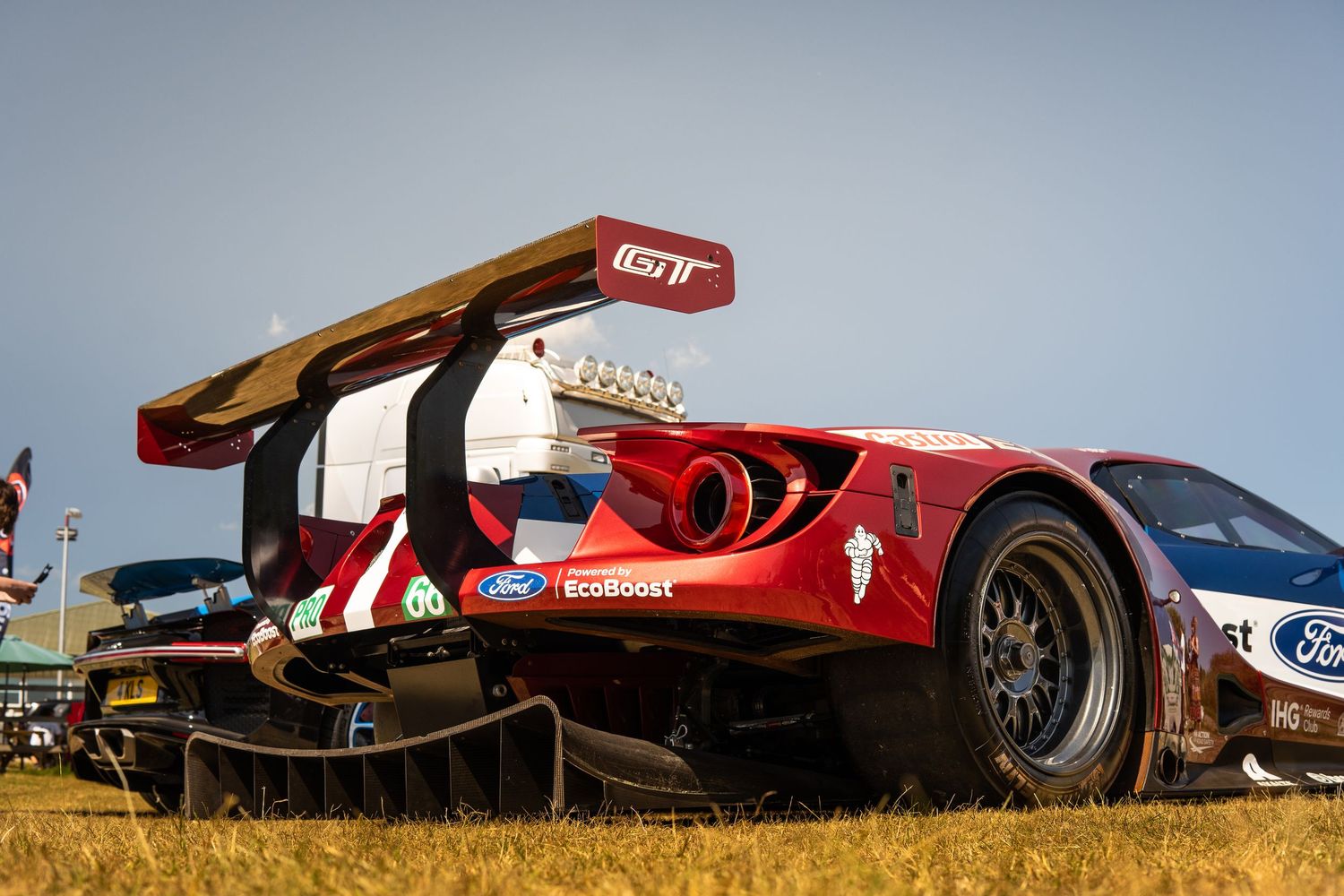 Red Ford GT race car with large rear wing at motorsport event on dry grass field.