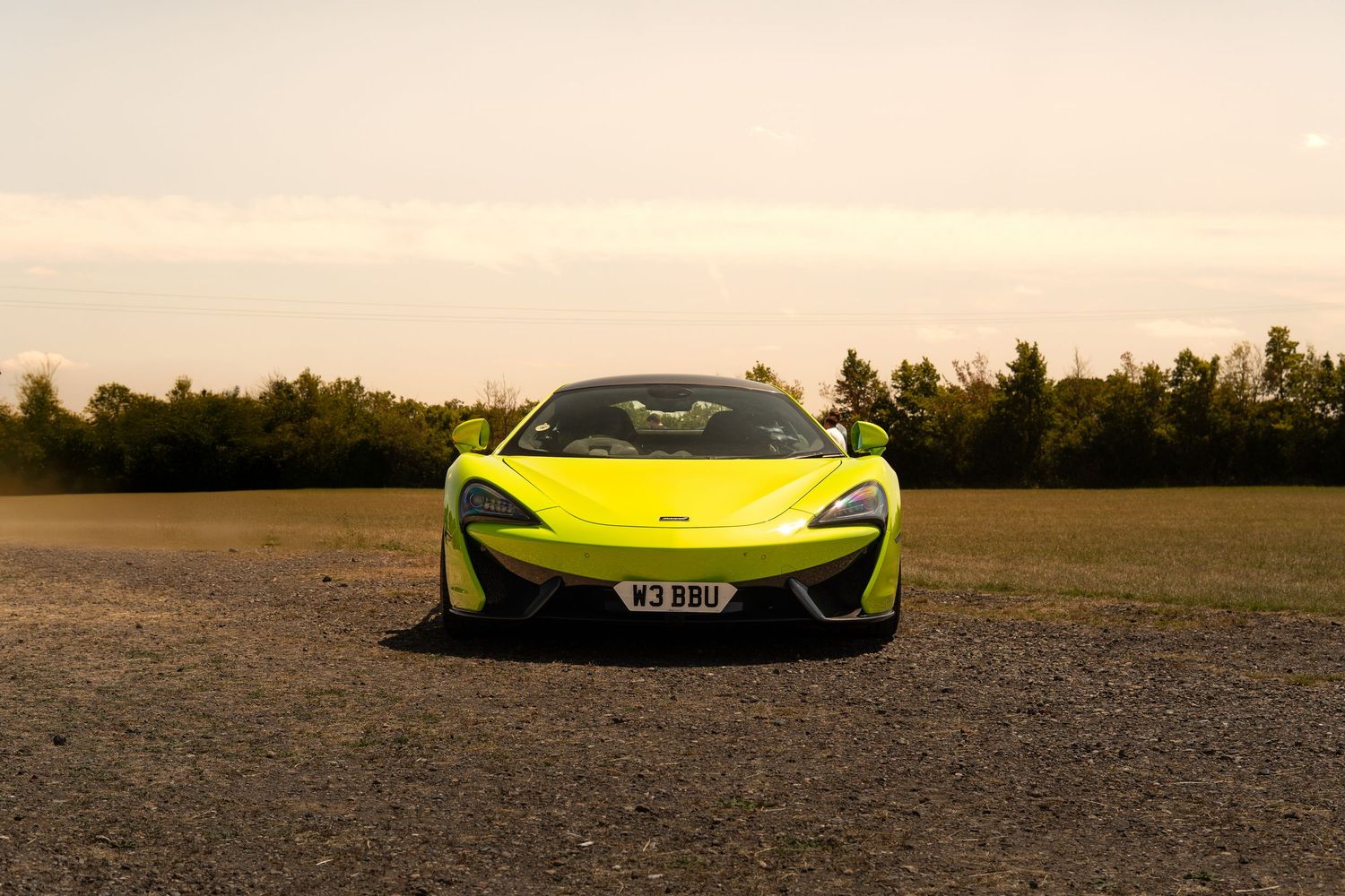 Bright yellow McLaren 570S supercar parked on gravel road at golden hour with trees in background.
