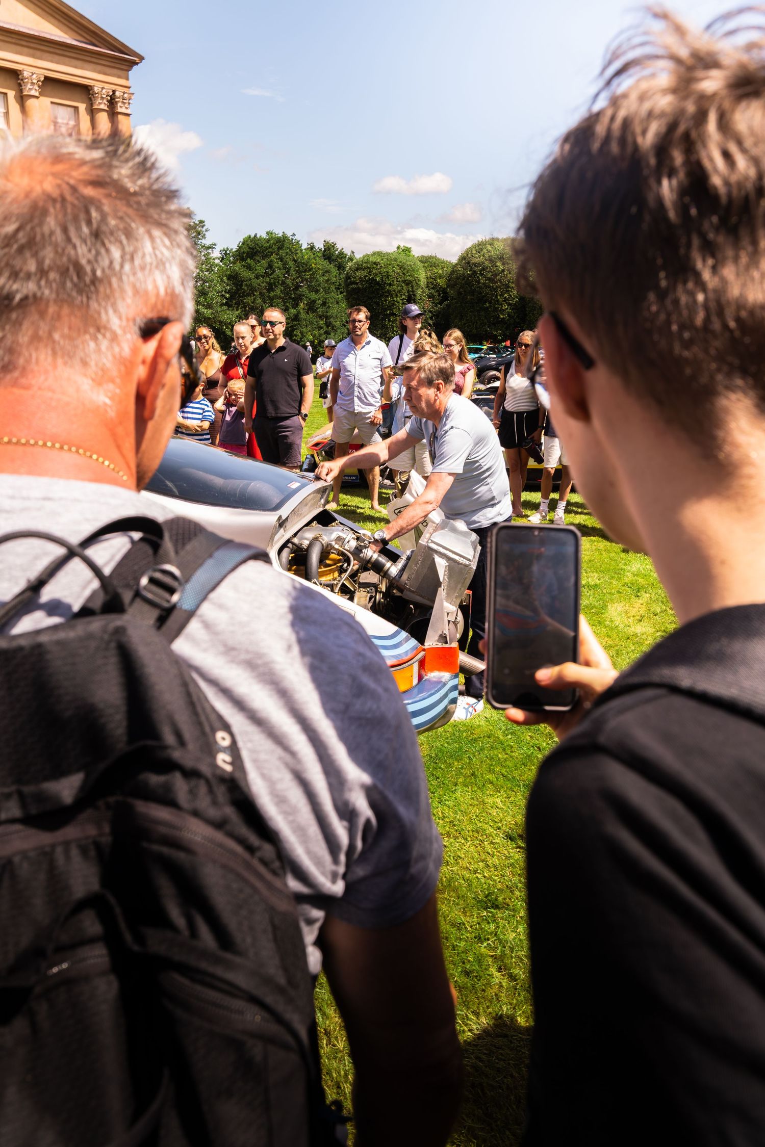 Crowd of people gathered outdoors on grass watching a demonstration near a classic building on a sunny day.