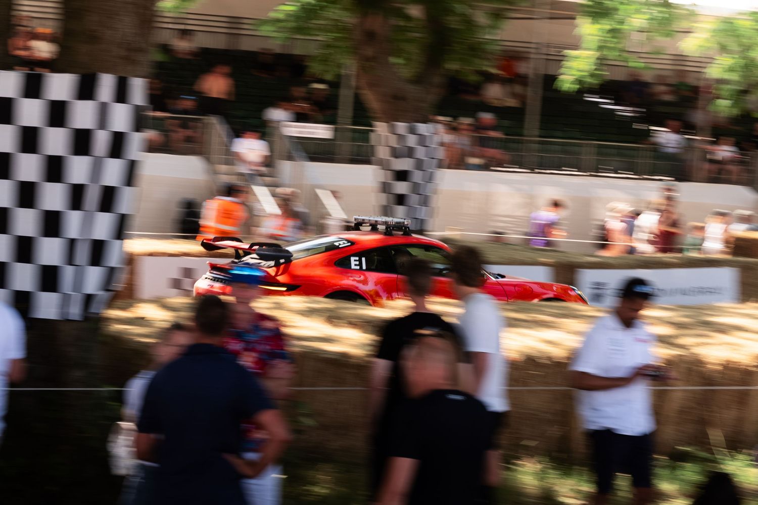 Red Formula 1 race car speeding past checkered flags at an outdoor motorsport hillclimb event with spectators watching.