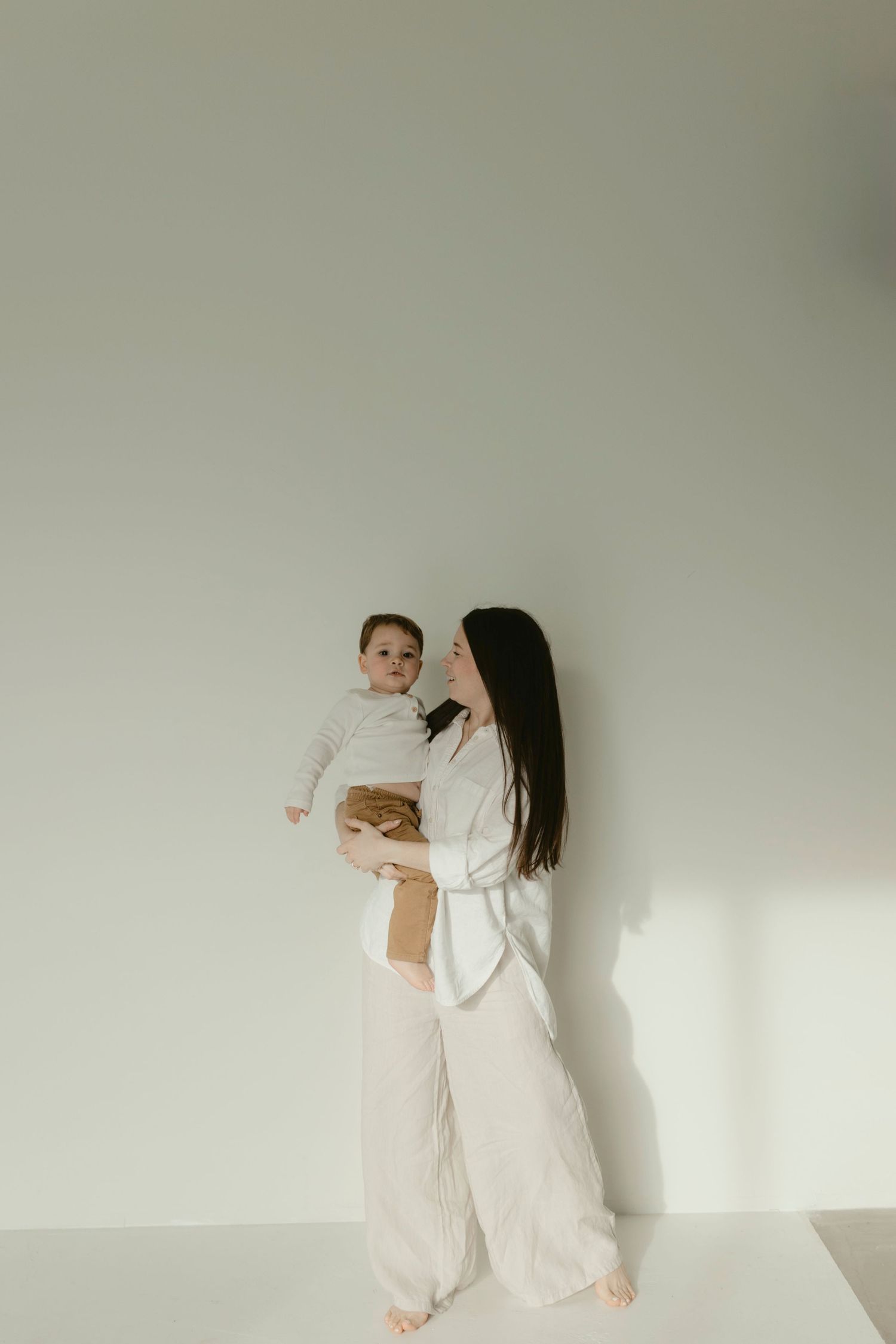 A mom and toddler in white clothing dance together against a plain wall, creating a minimalist and ethereal scene.