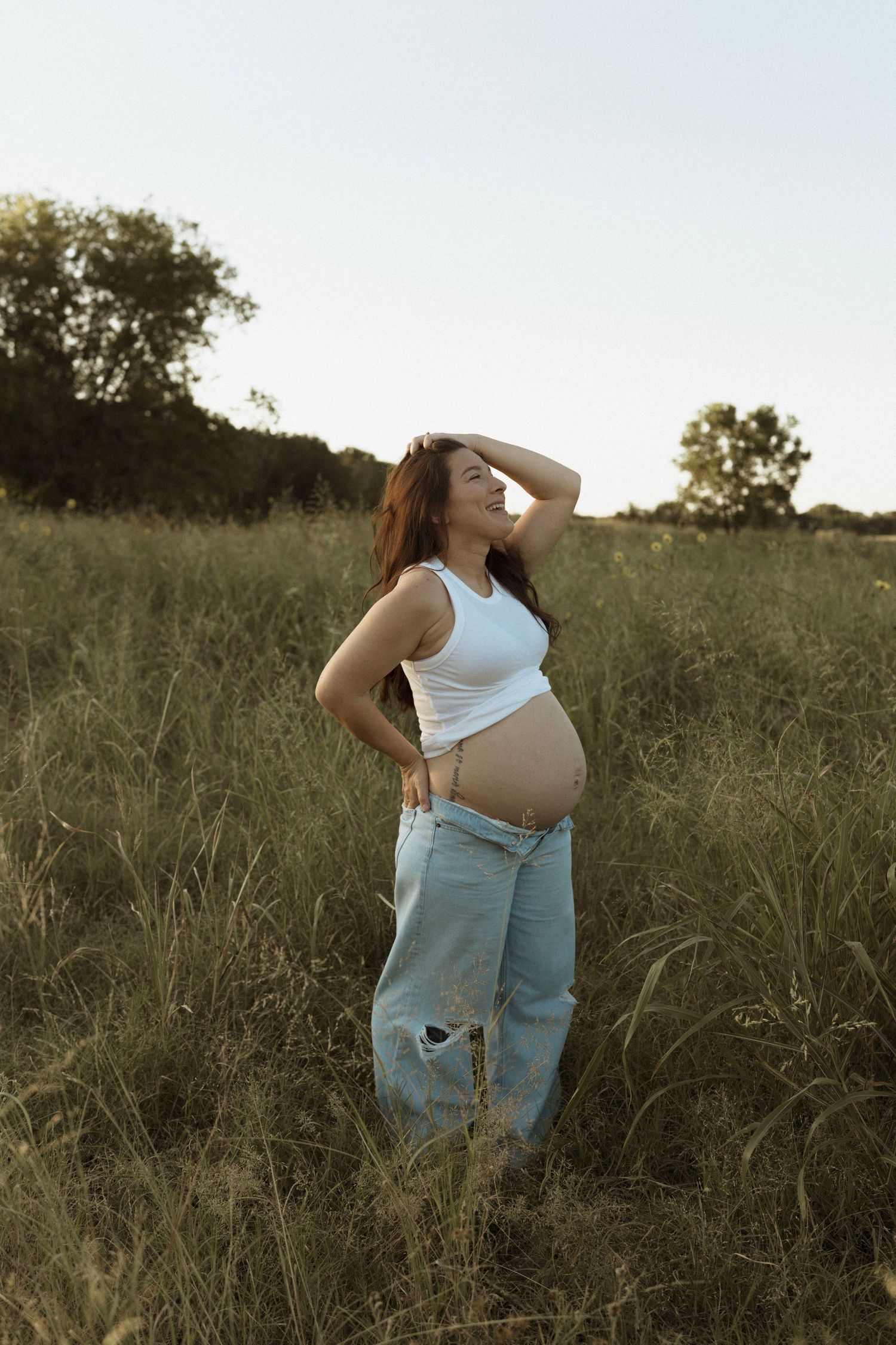 A pregnant woman in ripped jeans and white tank top poses in a grassy field at sunset.