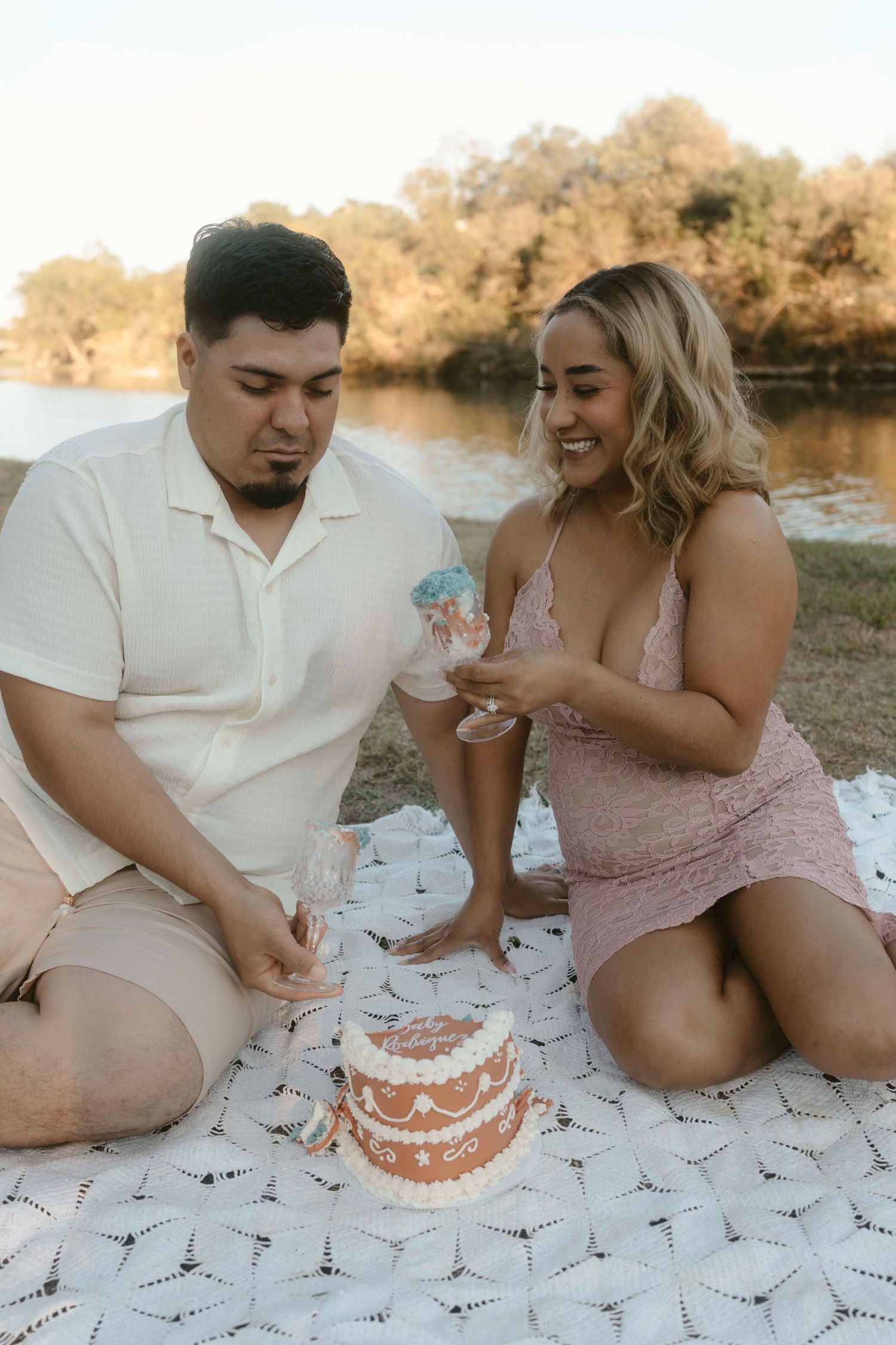 A couple sits on a blanket outdoors sharing cake during a casual picnic celebration at sunset.