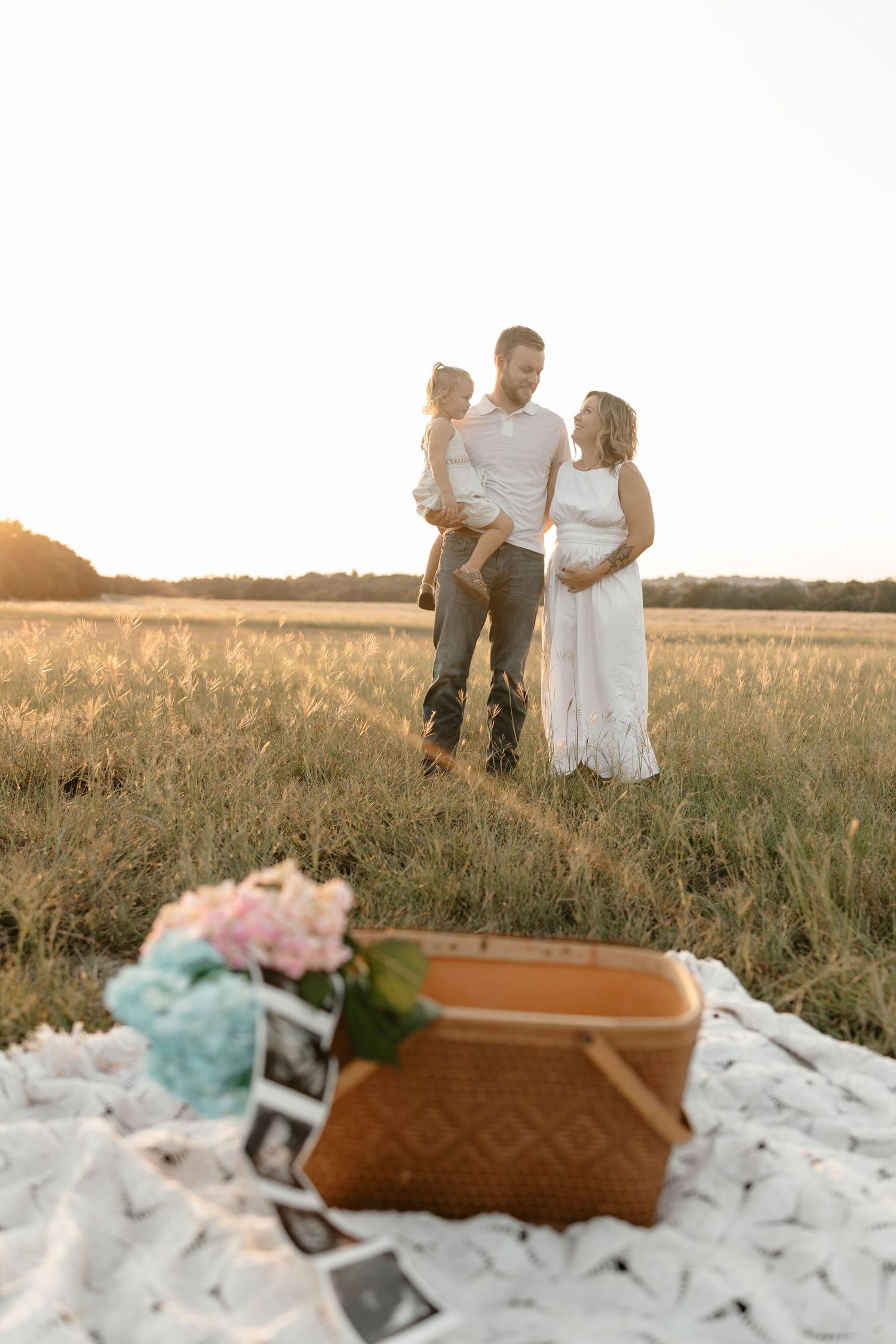 A couple stands together in a golden wheat field at sunset with a pregnancy announcement basket in the foreground.
