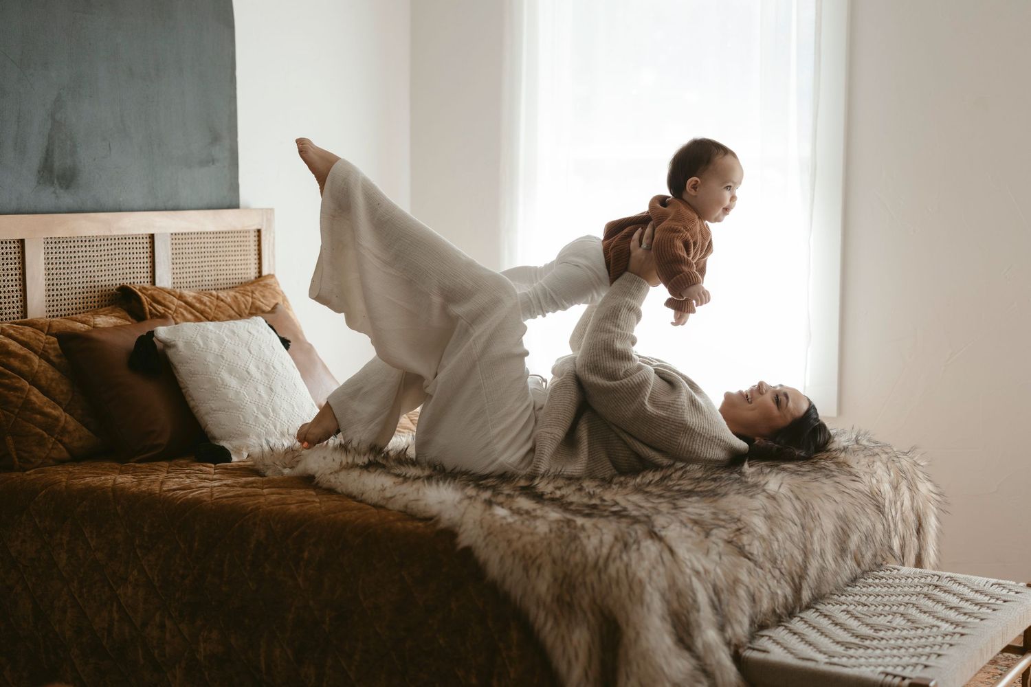 Playful family photo moment on bed with white linens and brown pillows, holding baby in the air.