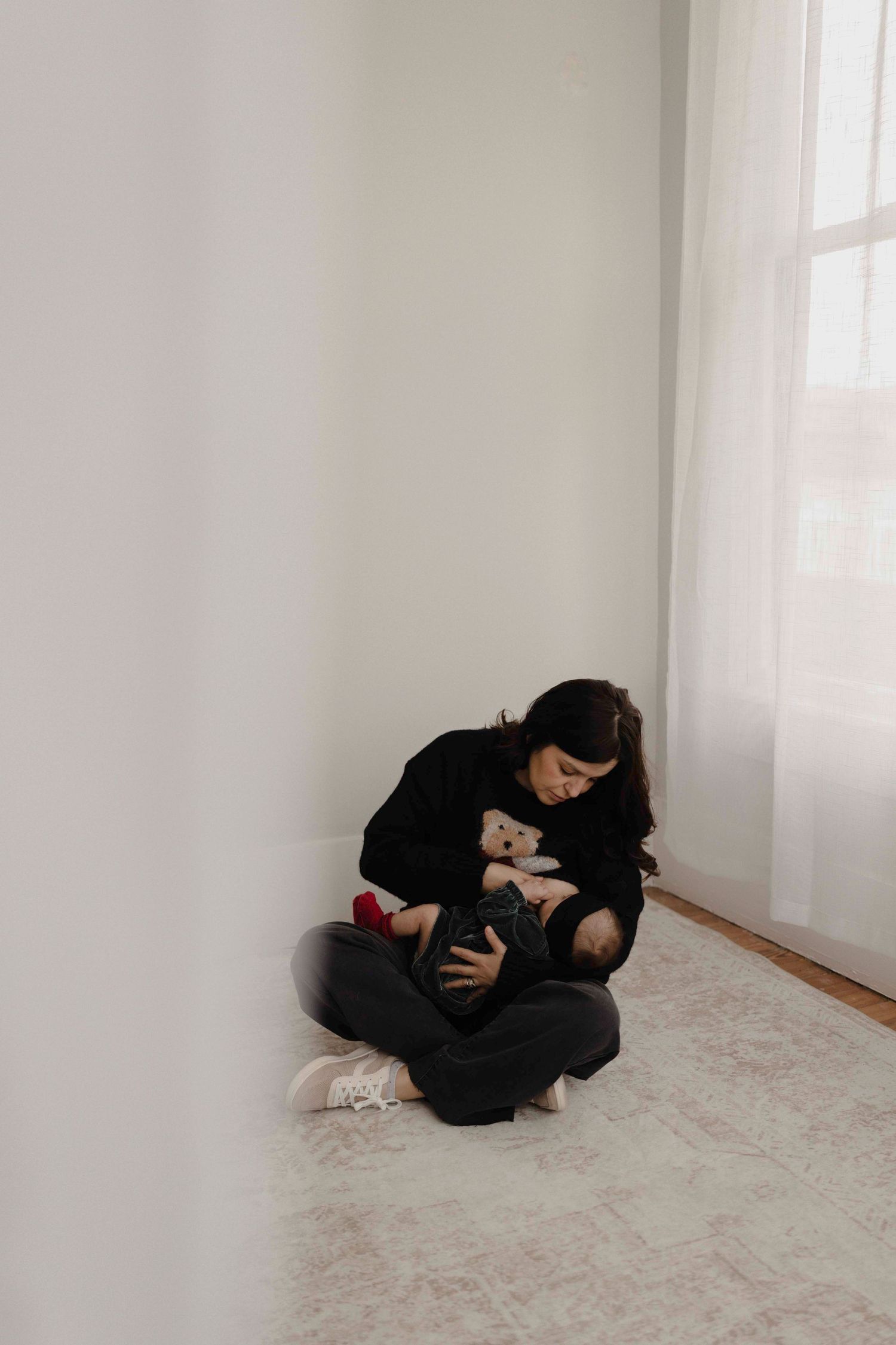 A person wearing black clothing curls up in fetal position on a light colored carpet in a bright minimalist room.