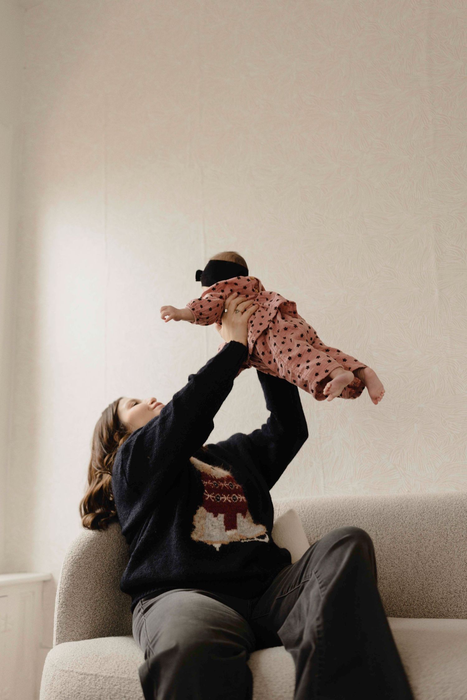Person in black sweater lying on couch playfully lifting baby wearing pink knit outfit in the air.