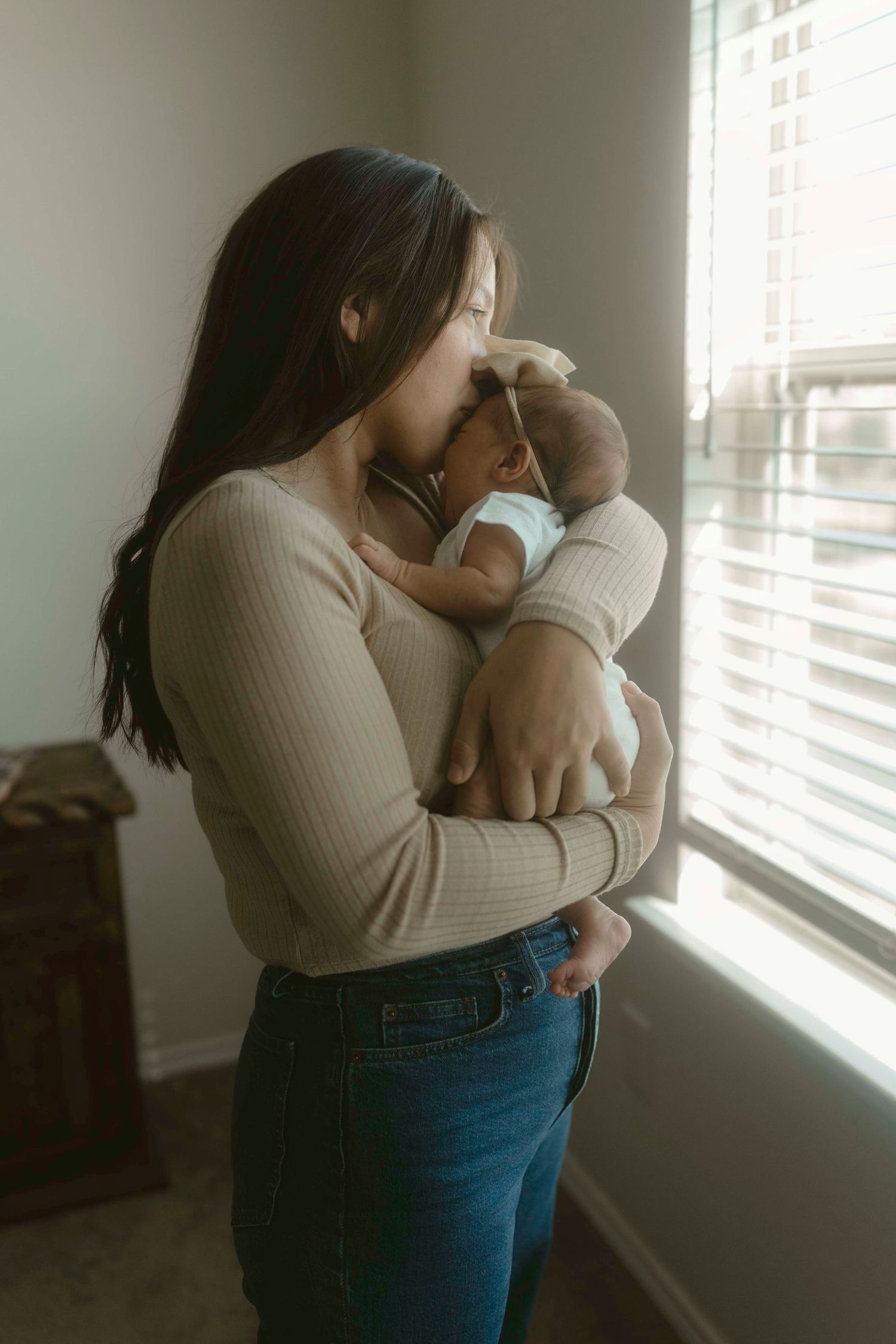A mother in a cream sweater and jeans stands by a window, holding and kissing her newborn baby