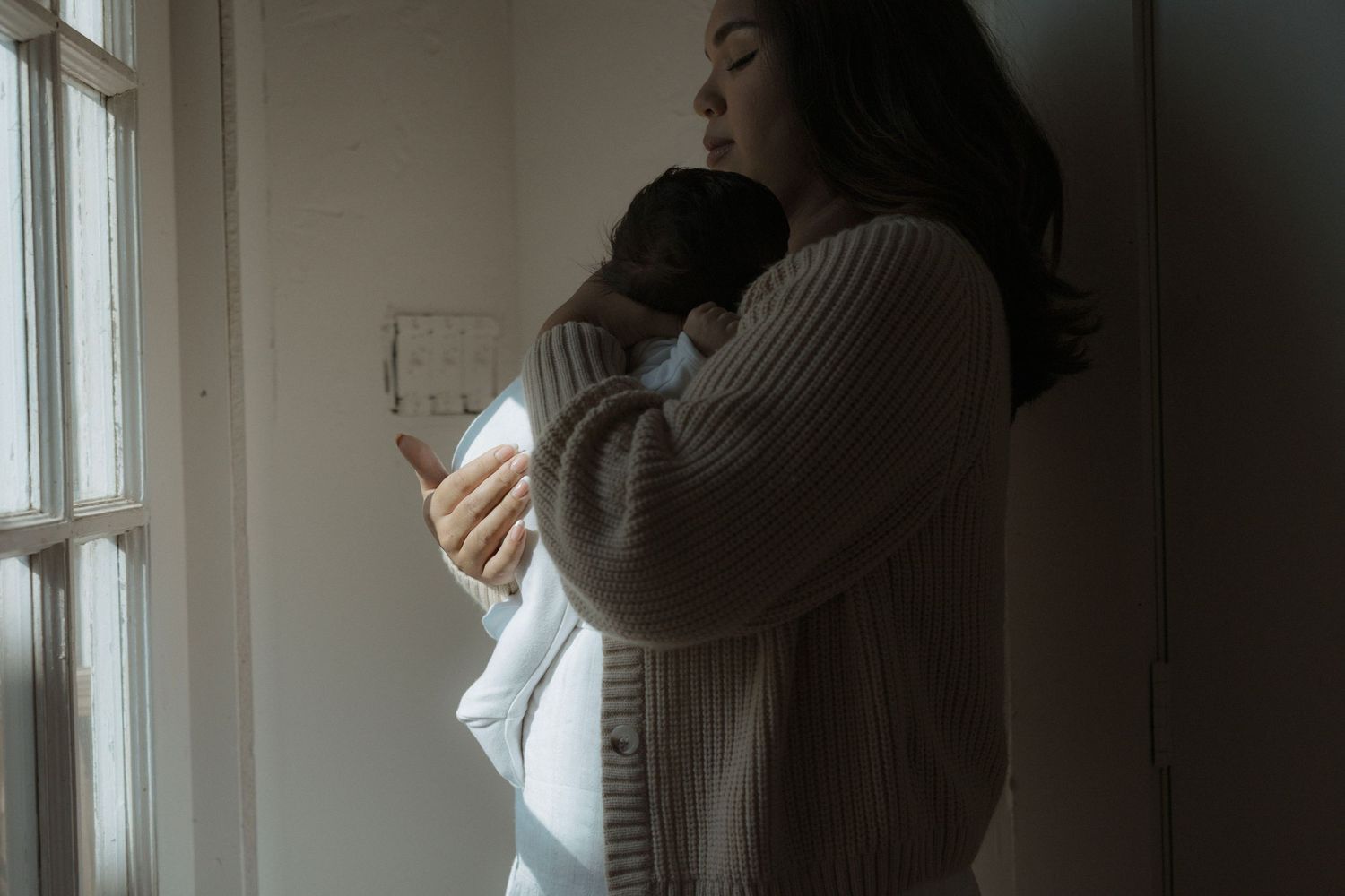 Cozy newborn photo of mother holding baby while looking out the window of her home.