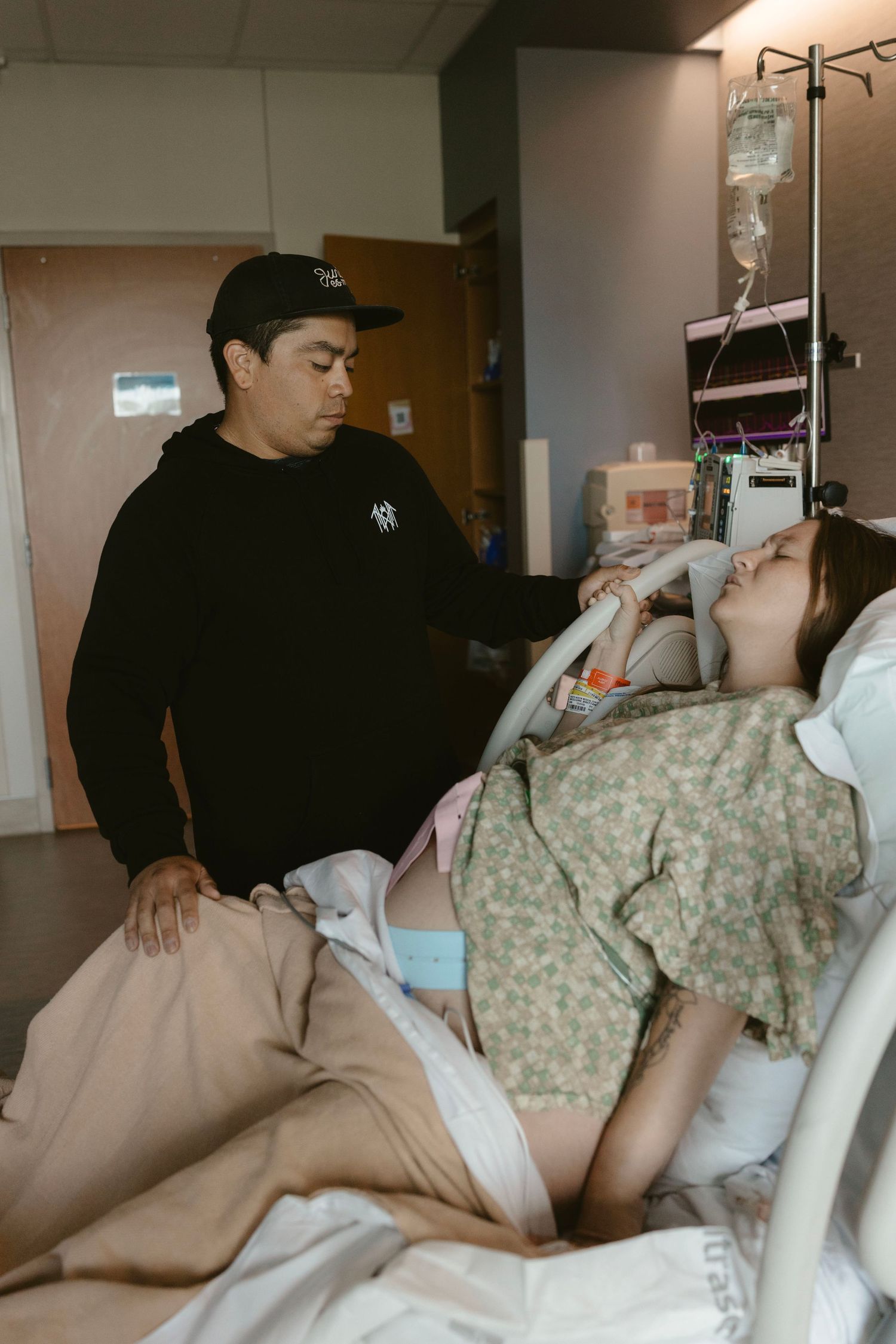 A person lies in a hospital bed wearing a medical gown while another person sits nearby in dark clothing.