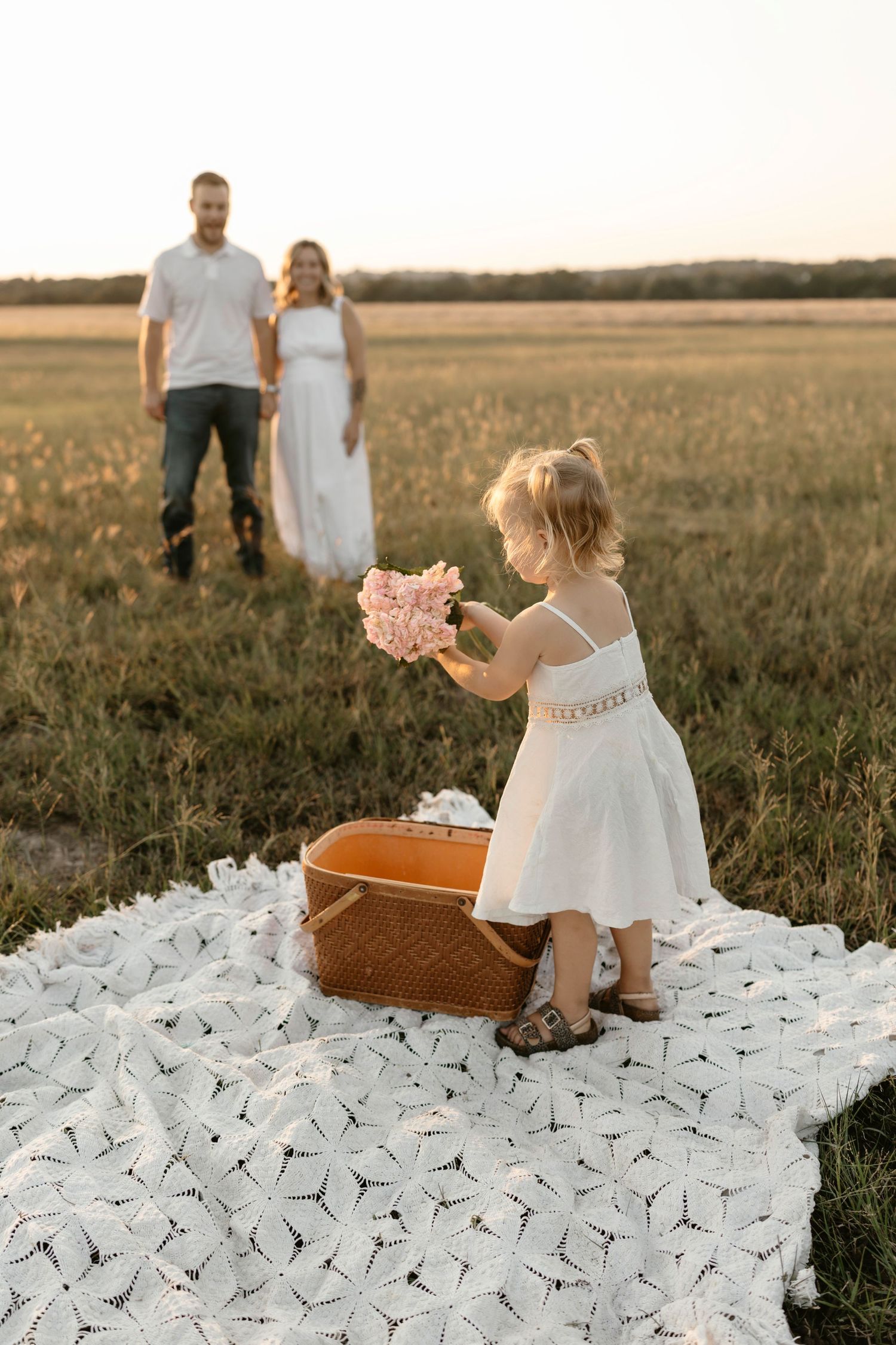 A couple stands in a golden field at sunset with a white lace dress and rustic picnic blanket spread on the grass.