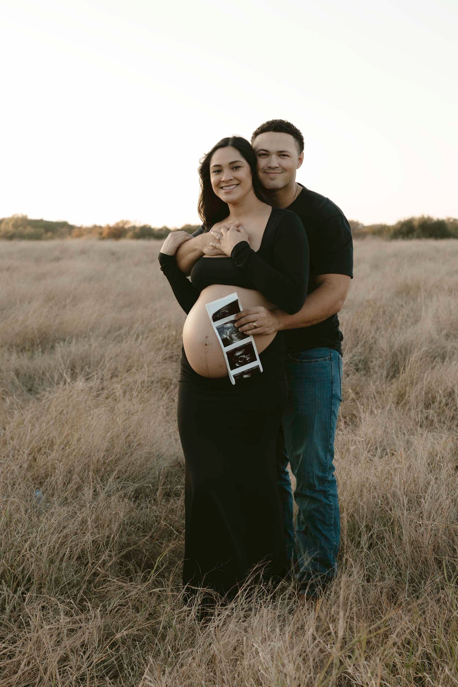 A maternity couple embraces in a golden field at sunset while holding an ultrasound photo.
