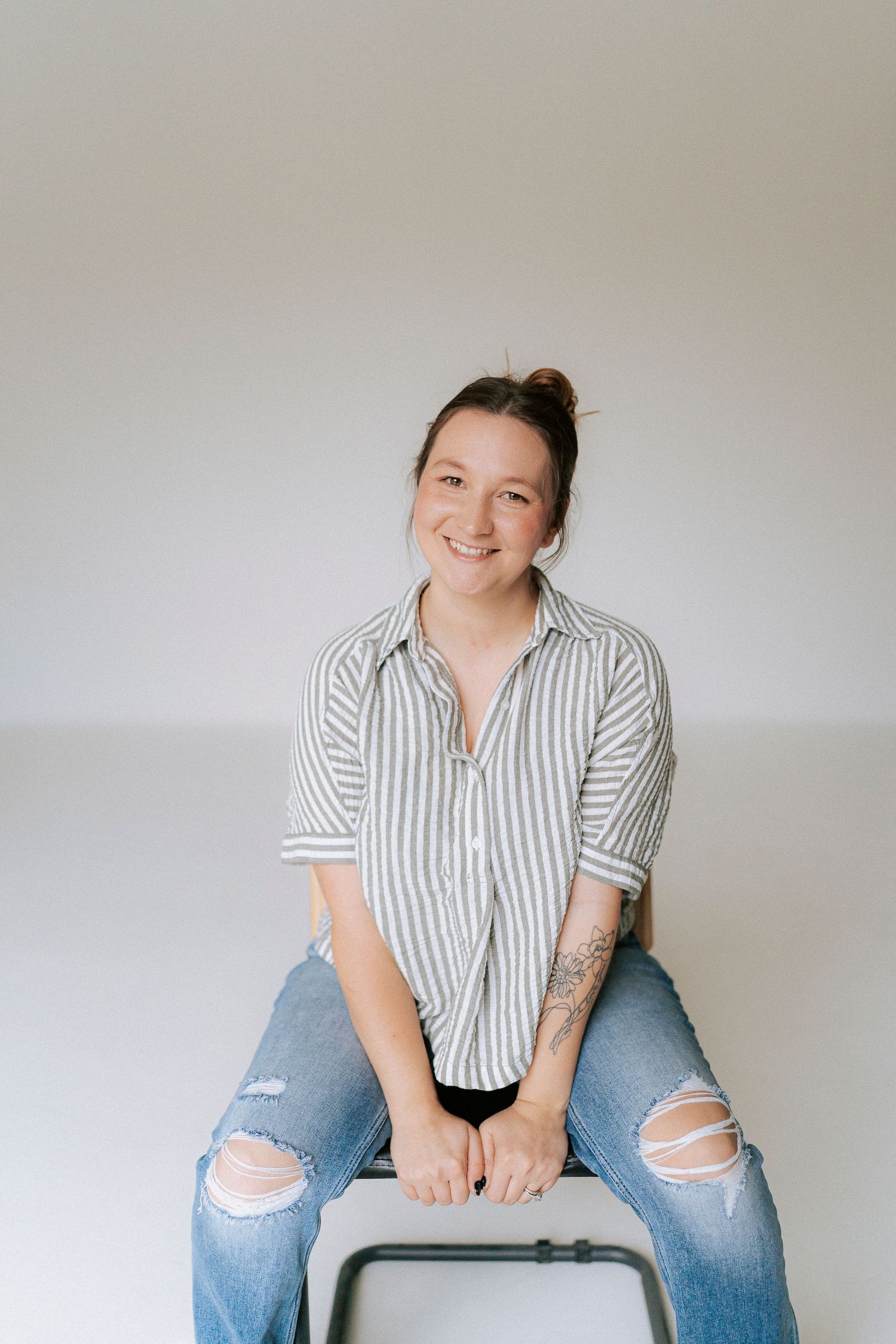 Person in striped button-down shirt and ripped jeans sits casually against white wall in lifestyle photoshoot series.