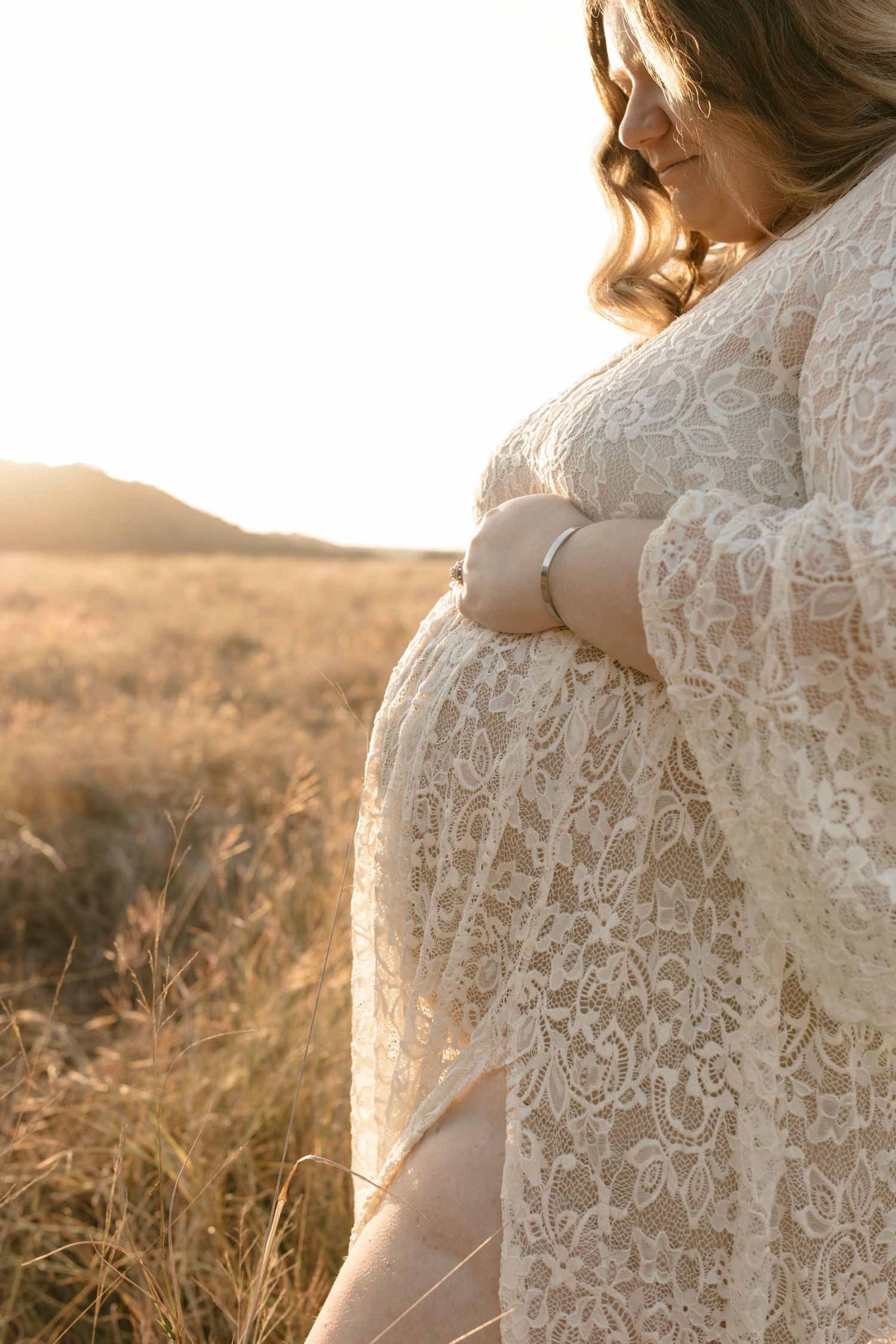 An expextant in a delicate lace dress stands in a golden field at sunset, with soft light illuminating the fabric details.
