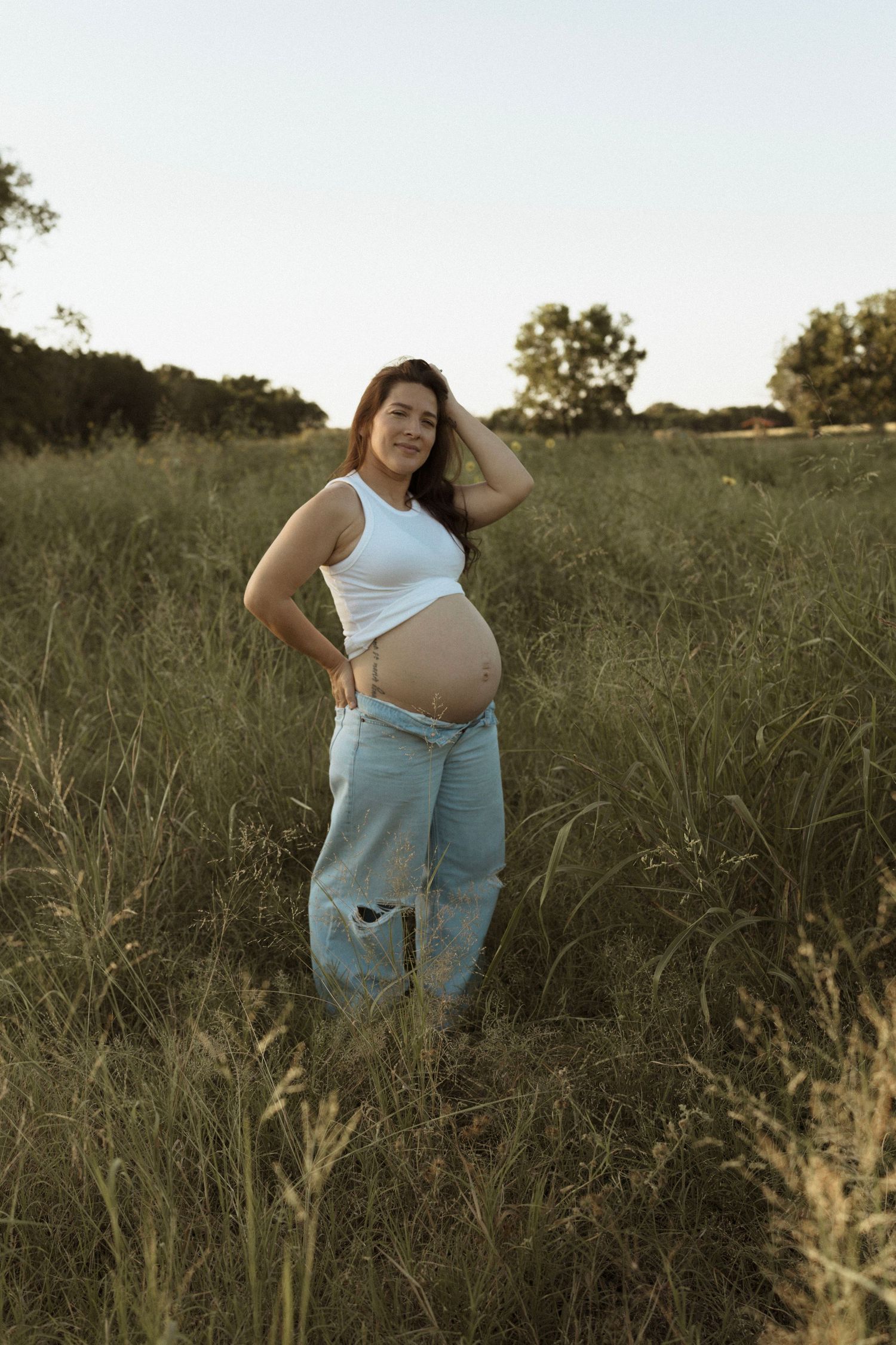 A mom poses in a grassy field at sunset wearing distressed jeans and a white crop top showing their pregnant belly.