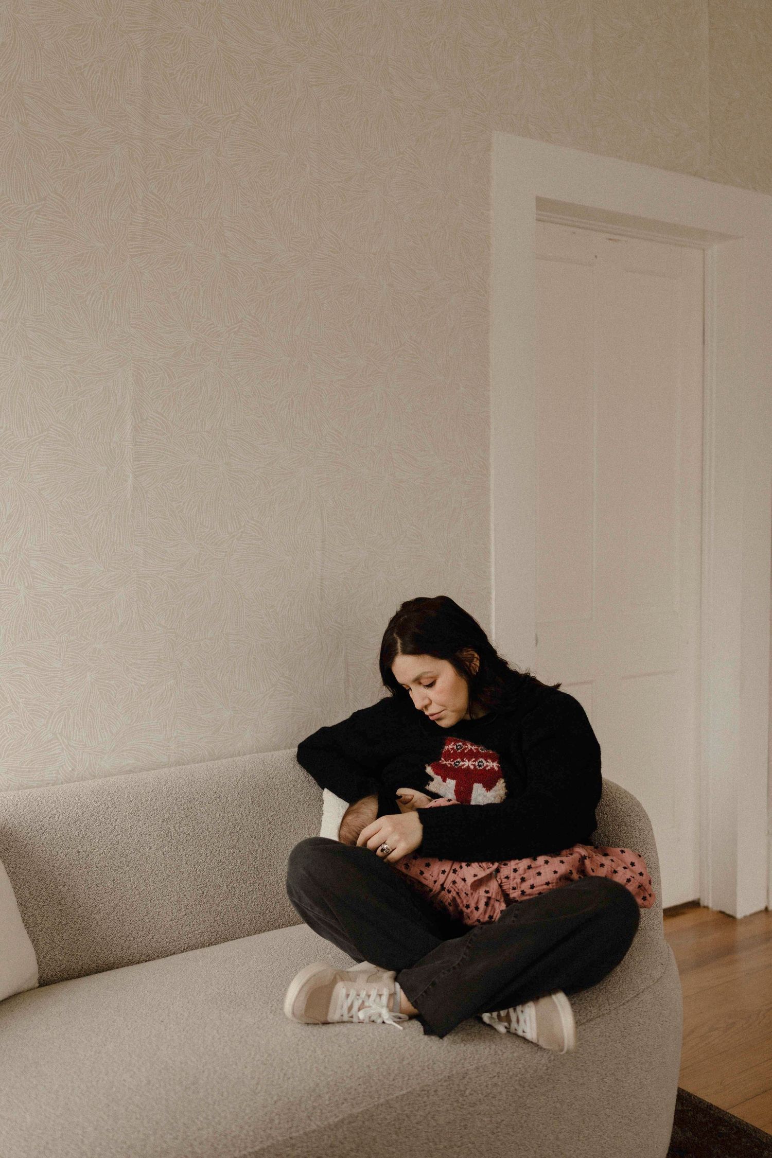 A person wearing black clothing curled up on a beige couch in a minimalist room with white walls.