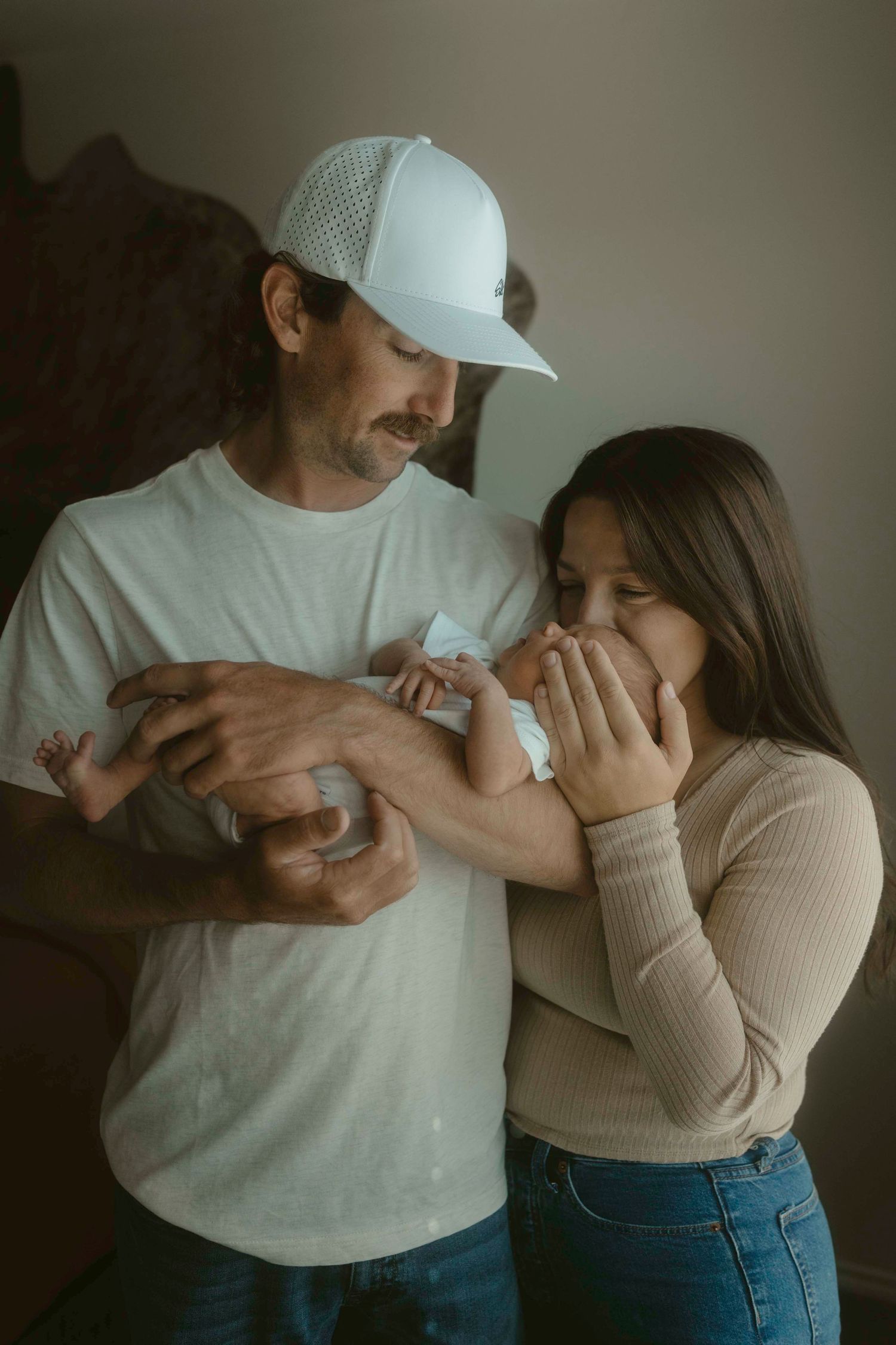 A couple shares an intimate moment while holding a baby, with one wearing a white baseball cap and t-shirt.