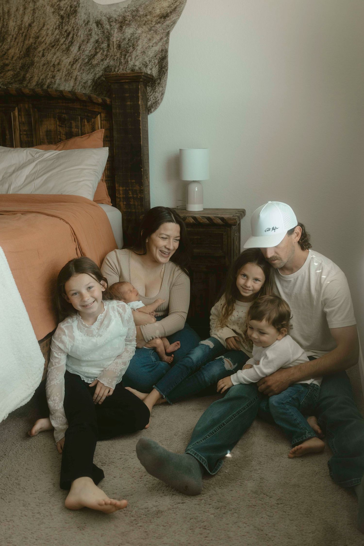 A big family of young kids sits together on the floor beside a bed, sharing a casual and intimate moment in a cozy bedroom setting.