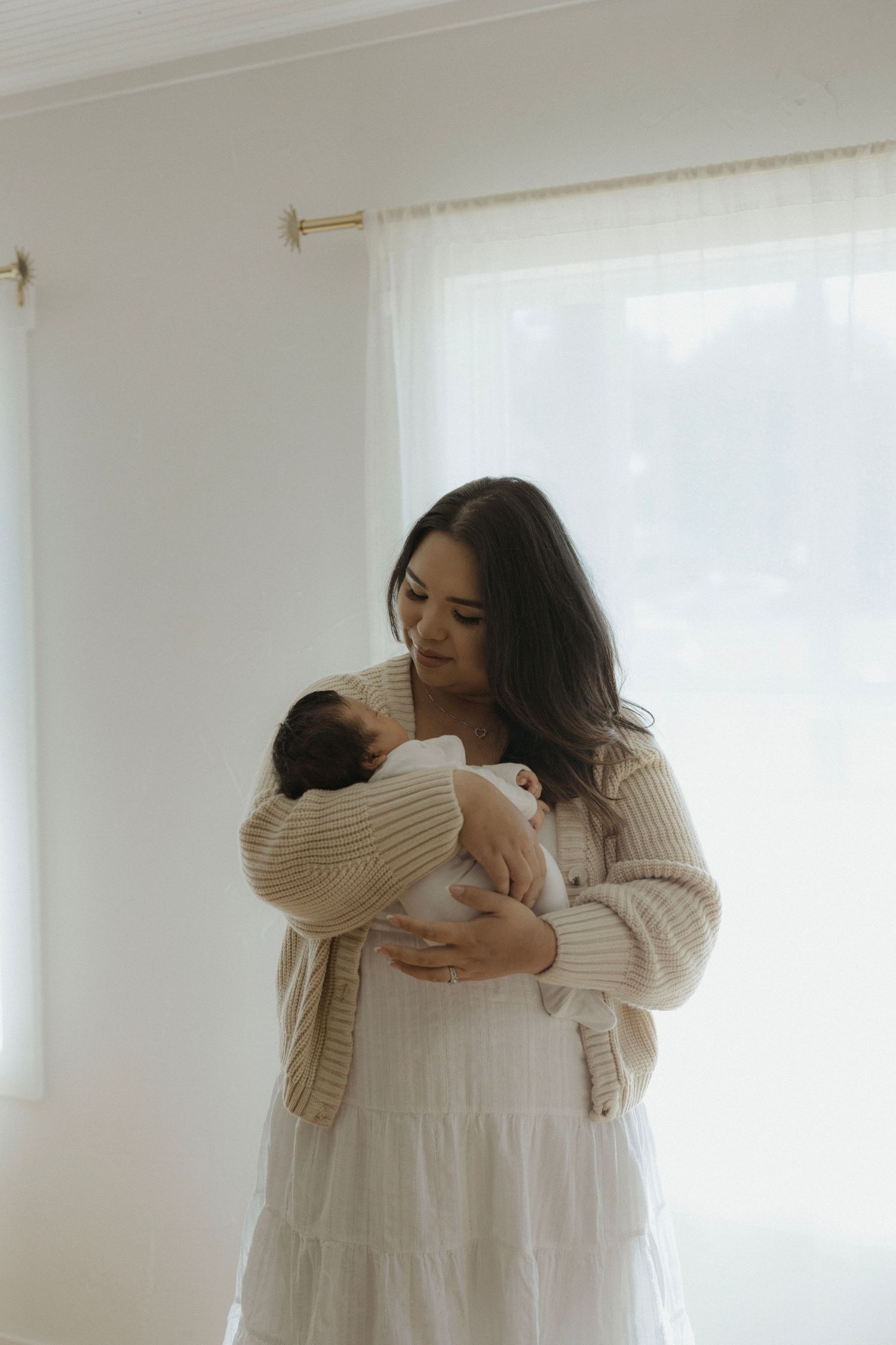 An Austin, TX mother in a cream sweater and white dress tenderly holds her newborn baby near a bright window during her photo session.