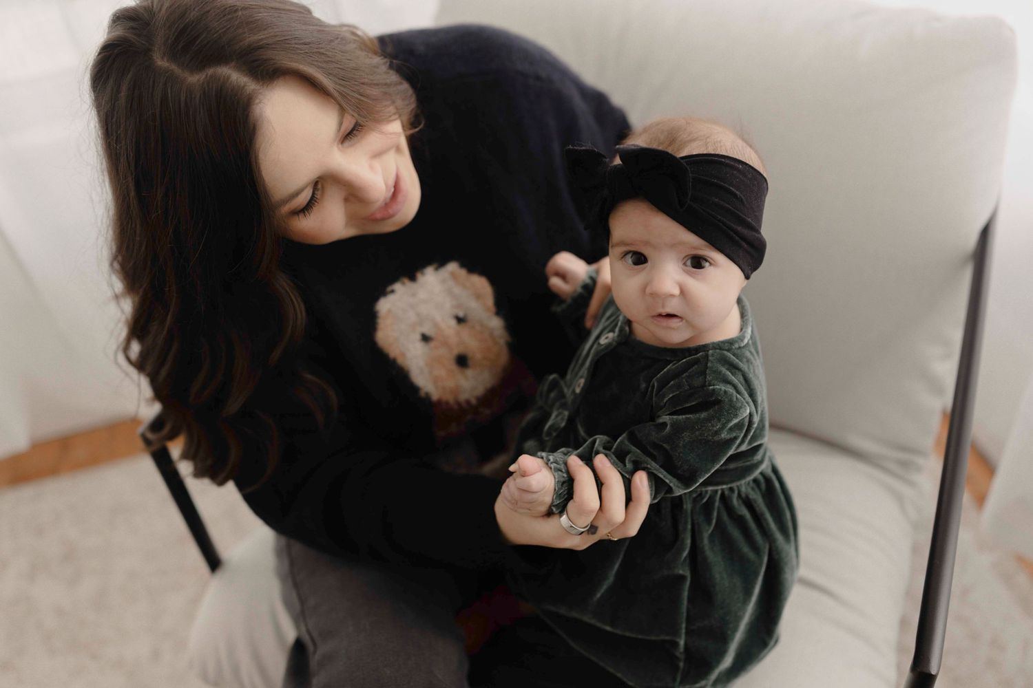 A person in a black sweater holds a baby wearing a dark green velvet outfit and black beanie on a light colored couch.