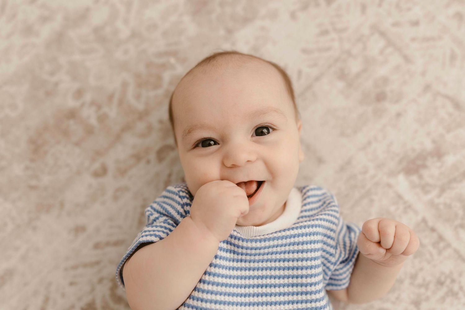 A baby in a blue and white striped outfit lies on a beige blanket smiling and playing with their hands.