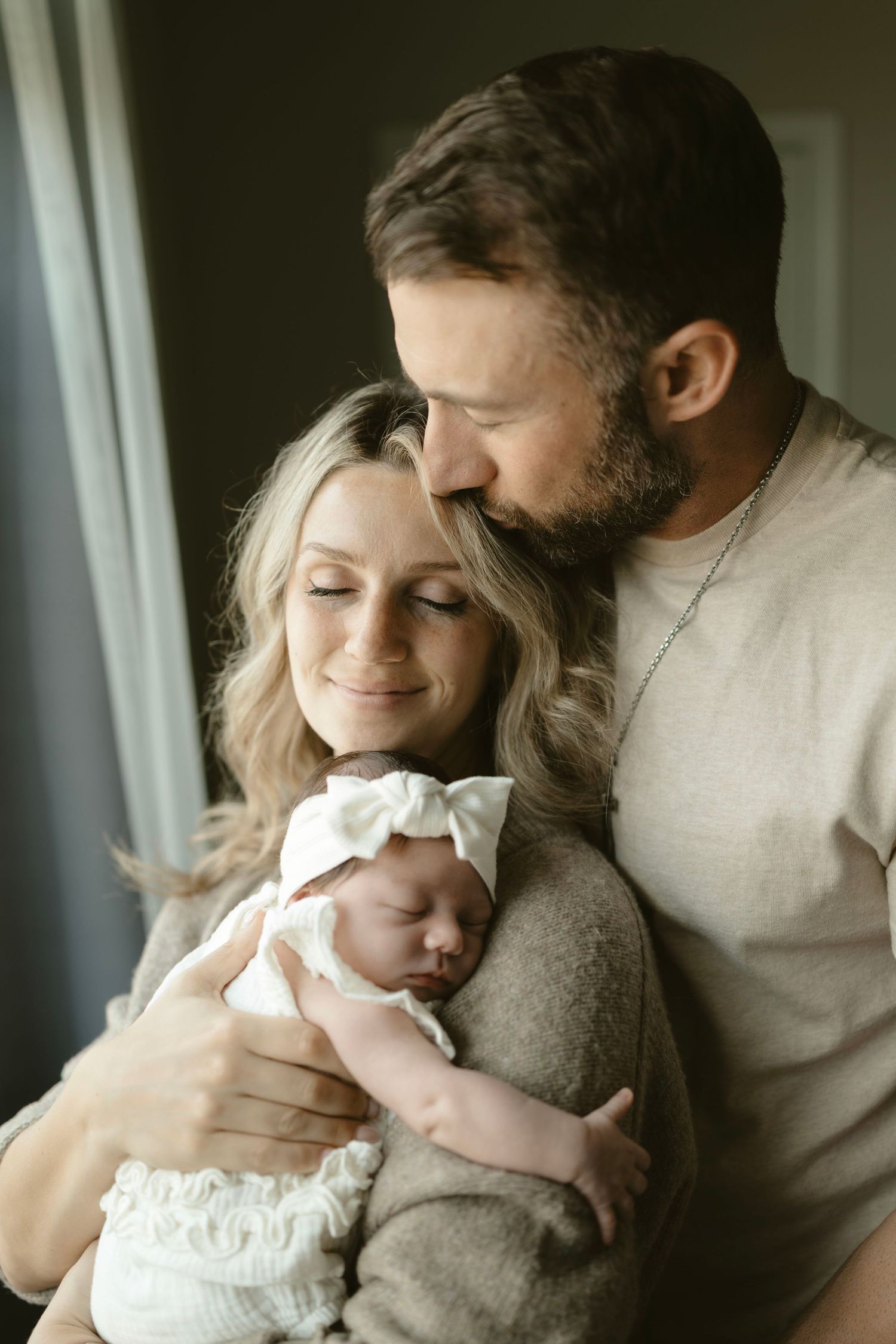 A couple shares an intimate moment while holding their sleeping newborn baby near a window in soft natural light.