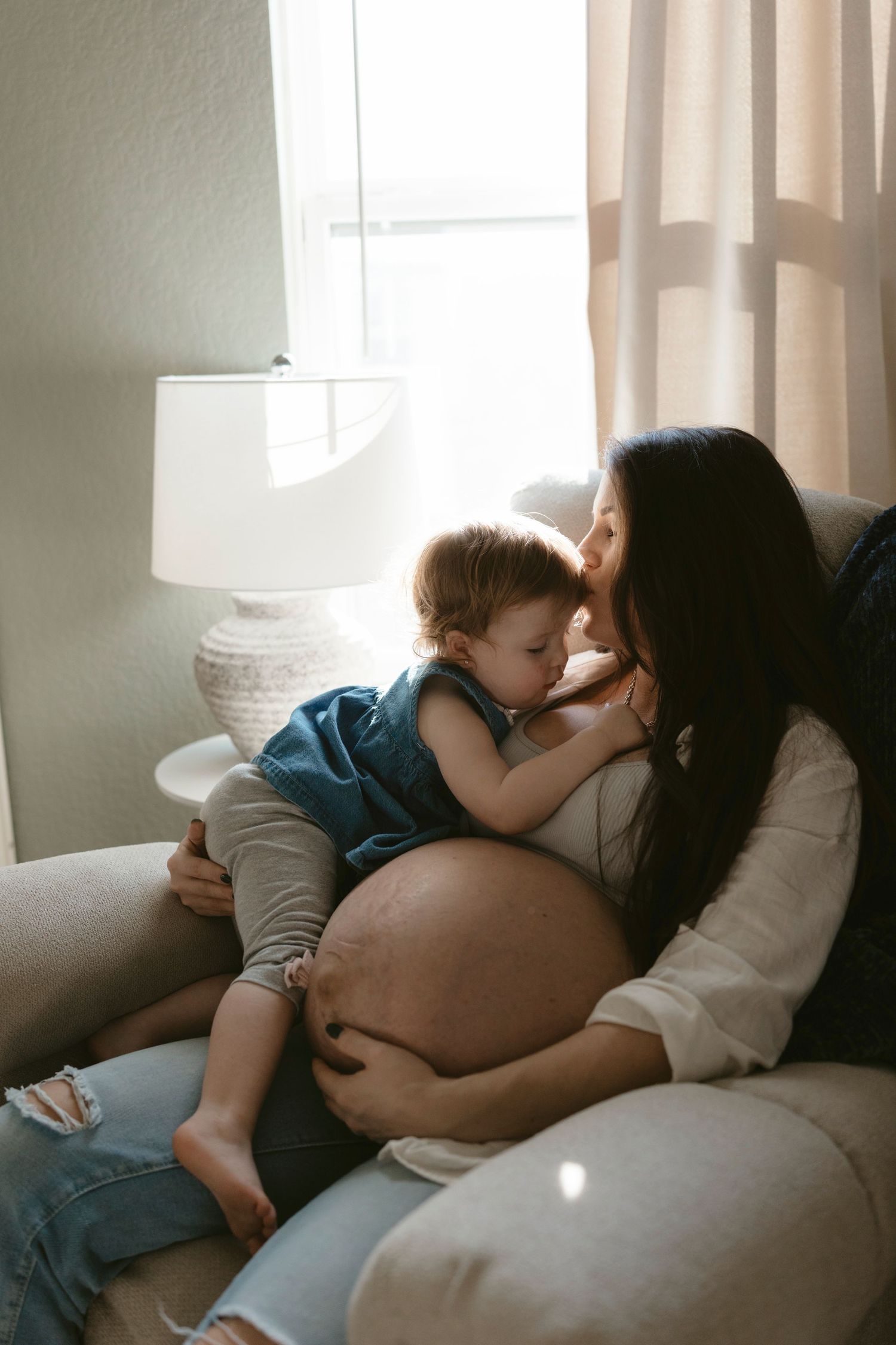 Intimate maternity moment with pregnant mom and her toddler cuddling on couch near window with soft natural light.