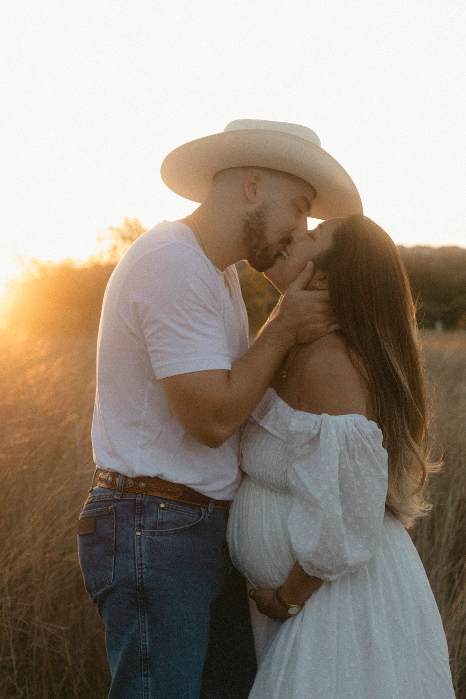 A romantic maternity couple embraces in a golden field at sunset, with soft lighting creating a dreamy silhouette.