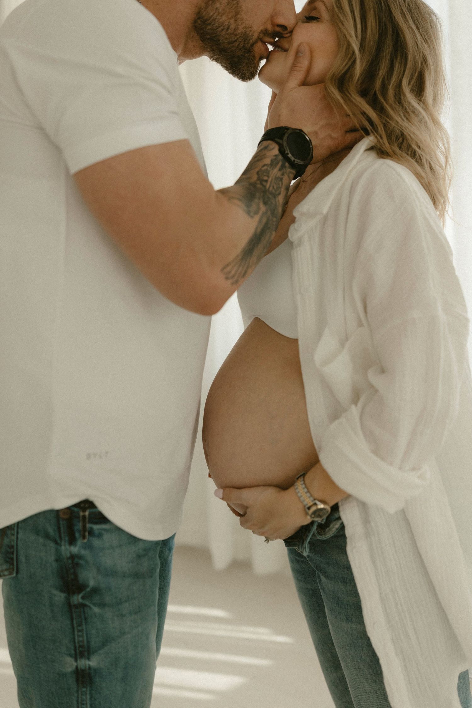 A couple in jeans and white shirts embraces intimately in a bright minimalist setting.